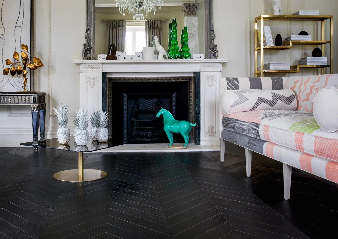 Sala de estar elegante con chimenea blanca decorada con adornos verdes y blancas, sofá con cojines de colores y patrón, mesa de centro negra con piñas plateadas, y estantería dorada con libros y objetos decorativos.