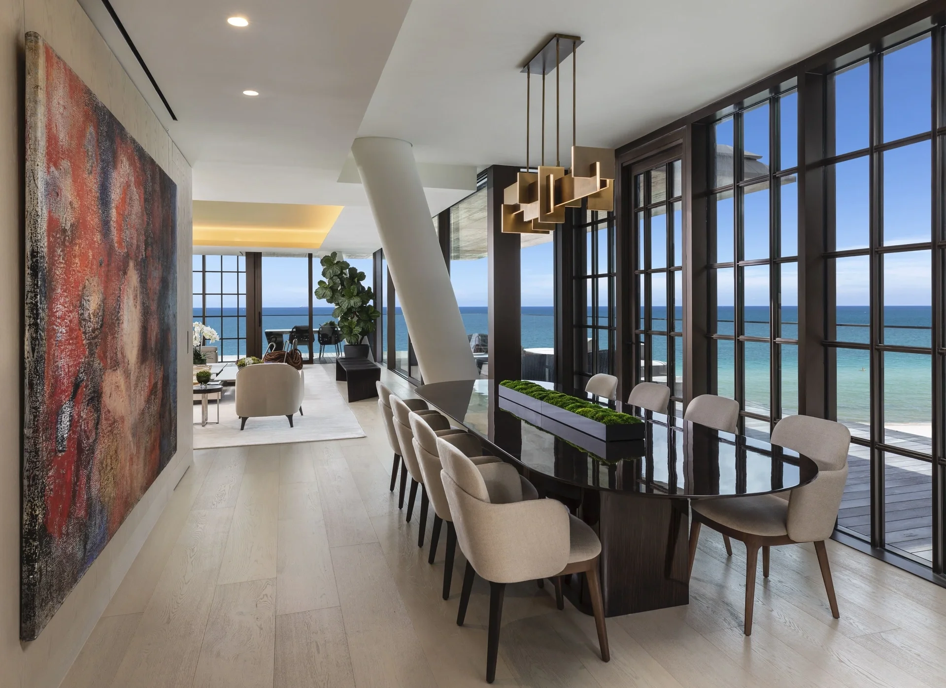 Interior de una sala moderna con vista al mar, mesa de comedor ovalada, sillas acolchonadas, gran ventanal. Decoración elegante con obra de arte y plantas.
