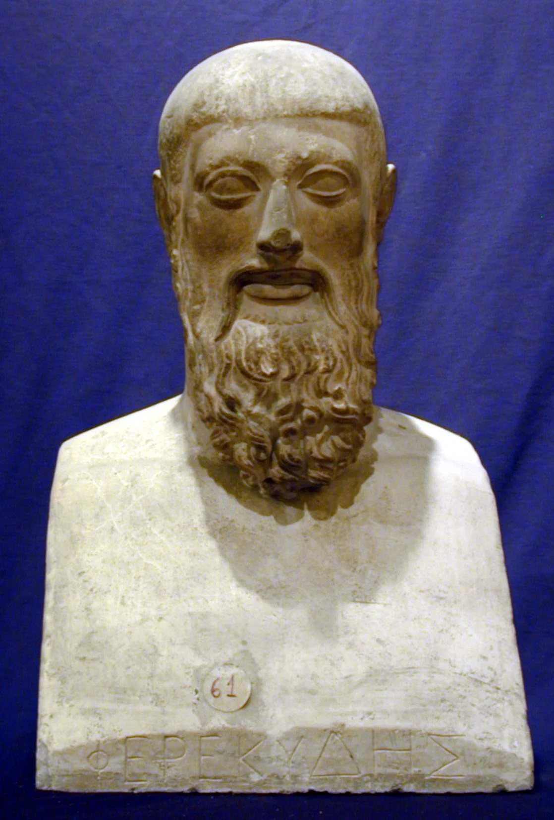 Busto de piedra de un hombre con barba y bigote, ojos cerrados, escrito en la base: "Ο ΠΕΚΥΛΑΔ" y un círculo con "61".