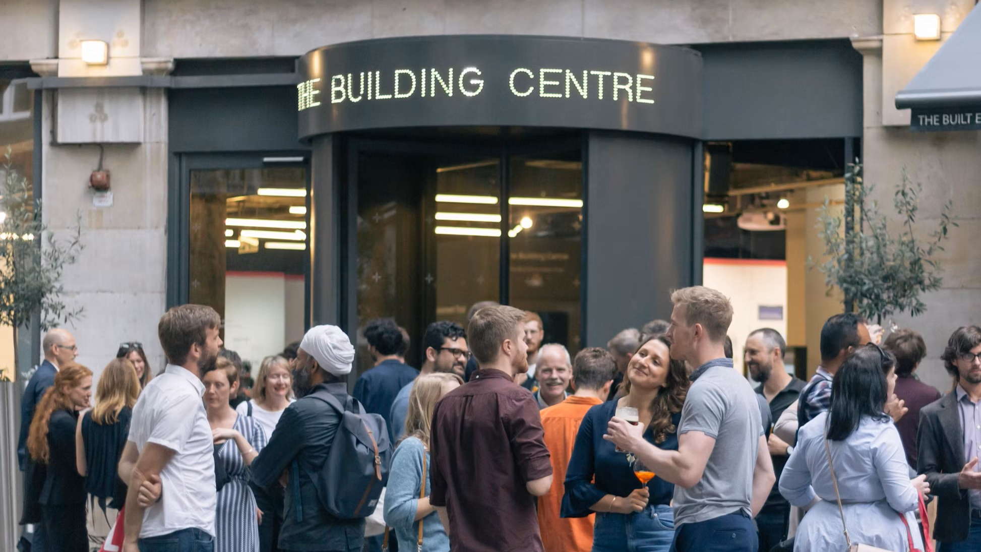 Gente reunida frente a un edificio con cartel que dice 'The Building Centre' en letras luminosas.