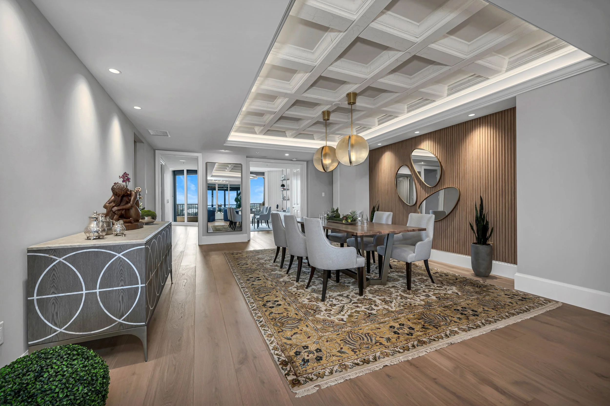 Sala de comedor moderna con mesa de madera y ocho sillas blancas, en un ambiente con pared de madera, espejos decorativos y plantas en macetas, con iluminación empotrada y lámparas colgantes de burbujas doradas.