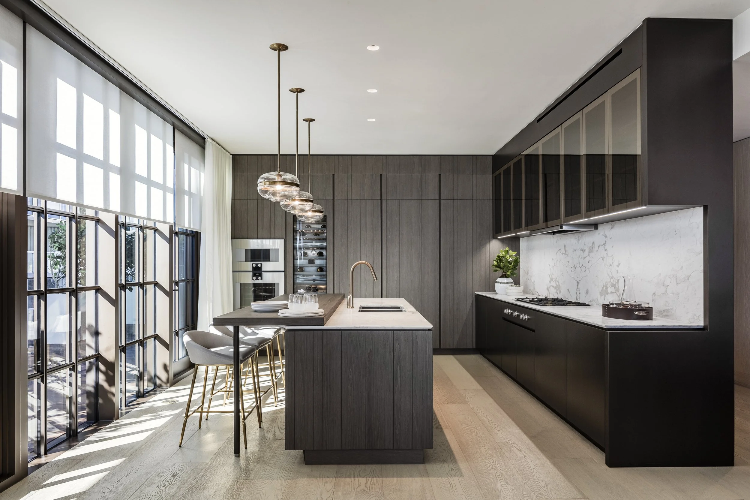 Cocina moderna con isla de mármol, gabinetes oscuros, ventanas grandes con persianas blancas, y lámparas colgantes de vidrio