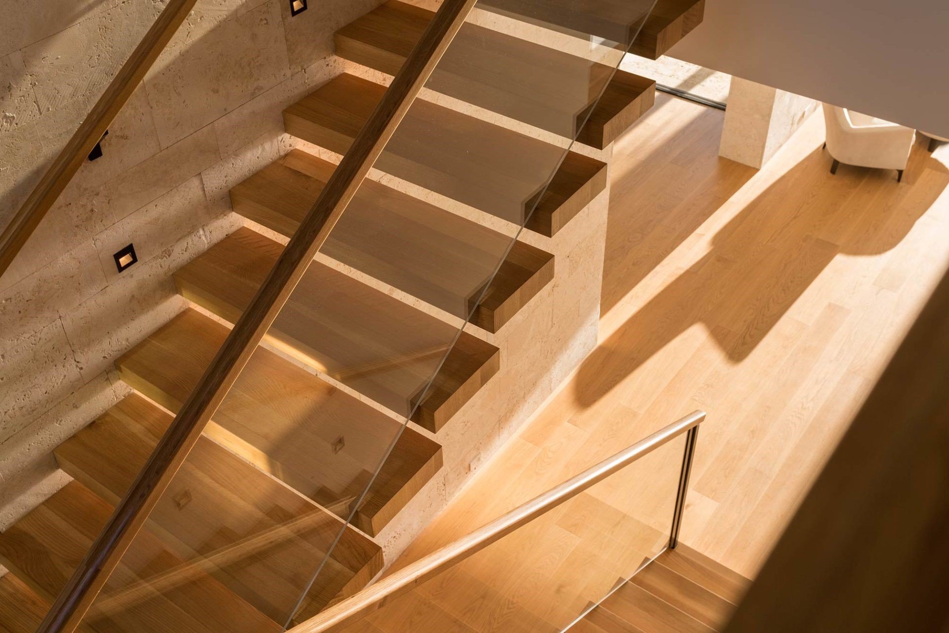 Vista aérea de una escalera moderna de madera con barandilla de metal y pared de piedra, con sombra proyectada en el piso de madera claro.