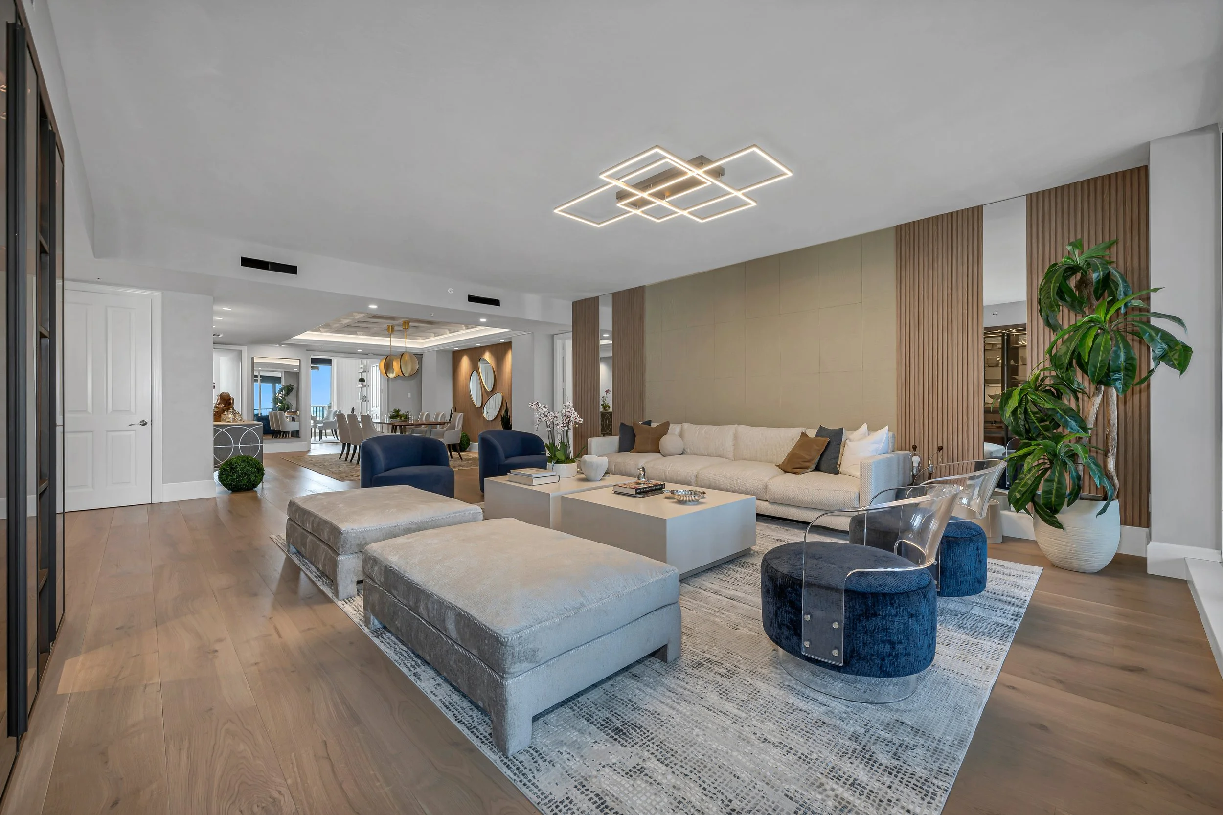 Sala de estar moderna con sofá beige, sillones transparentes, mesa de centro blanca y plantas decorativas en un ambiente luminoso y elegante.