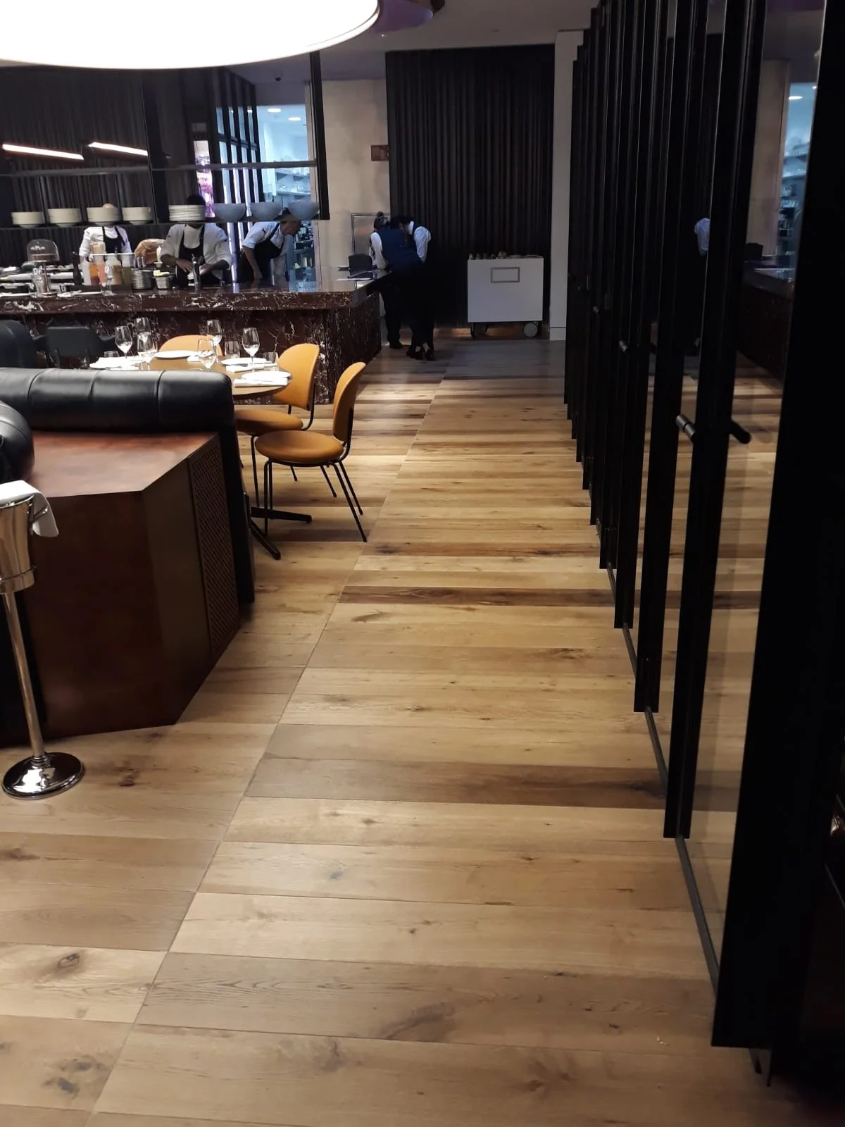 Interior de restaurante con mesas y sillas, área de cocina con personal preparando alimentos, piso de madera y paredes decoradas en tonos oscuros.