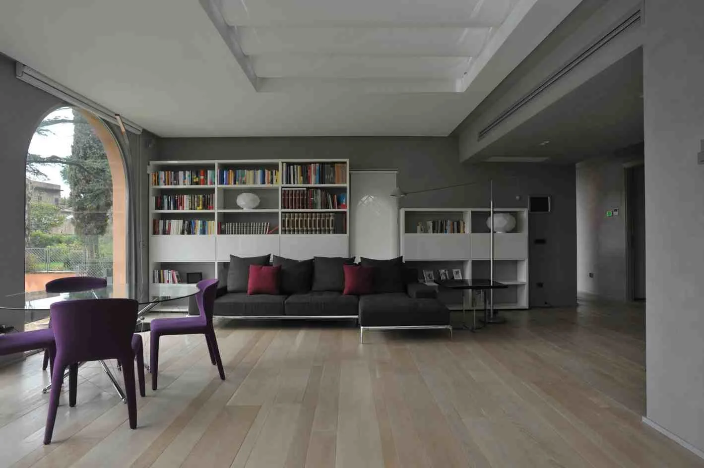 Sala moderna con librero, sofá negro con cojines burdeos, mesa de vidrio, sillas moradas, y puerta corrediza de vidrio con vista a un jardín