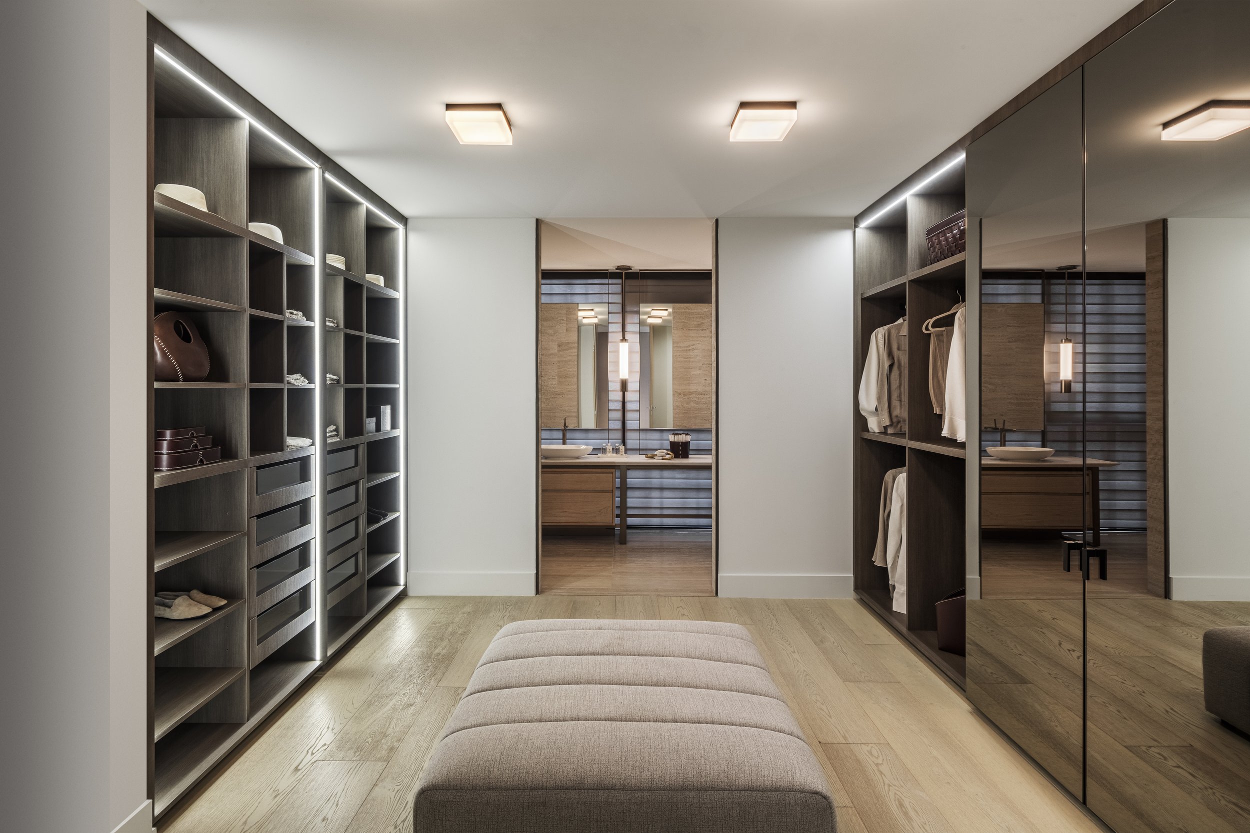 Vestidor moderno con armarios de madera, un banco acolchado en el centro, y un baño visible al fondo con lavabo y espejos.