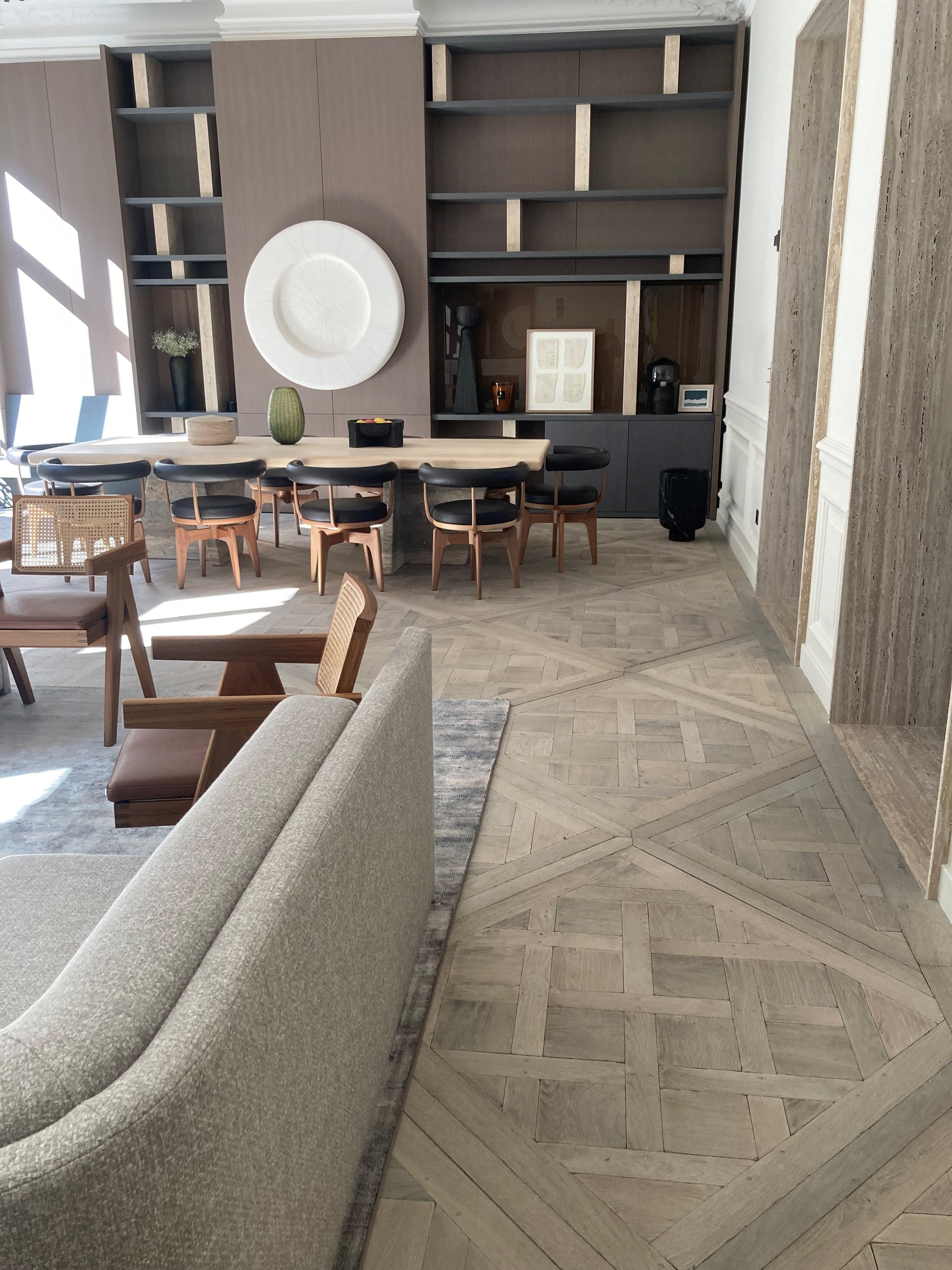 Sala de estar y comedor moderna con madera en tonos neutros, sillones, mesa extensible de madera, estantería con decoración y pisos de madera en patrón geométrico.