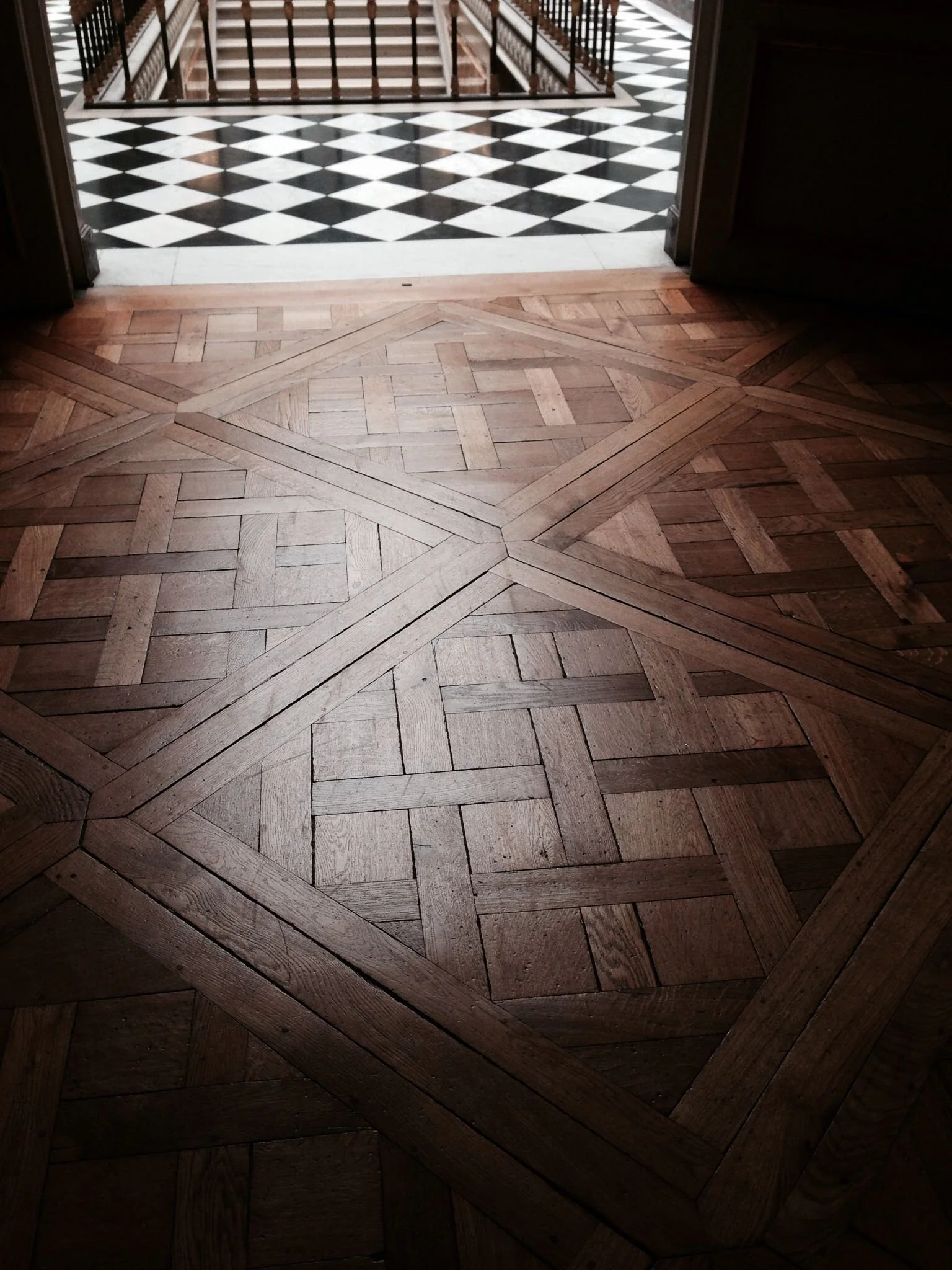 Suelo de madera con diseño geometric, y en el fondo un suelo de baldosas negras y blancas en patrón de rombos.