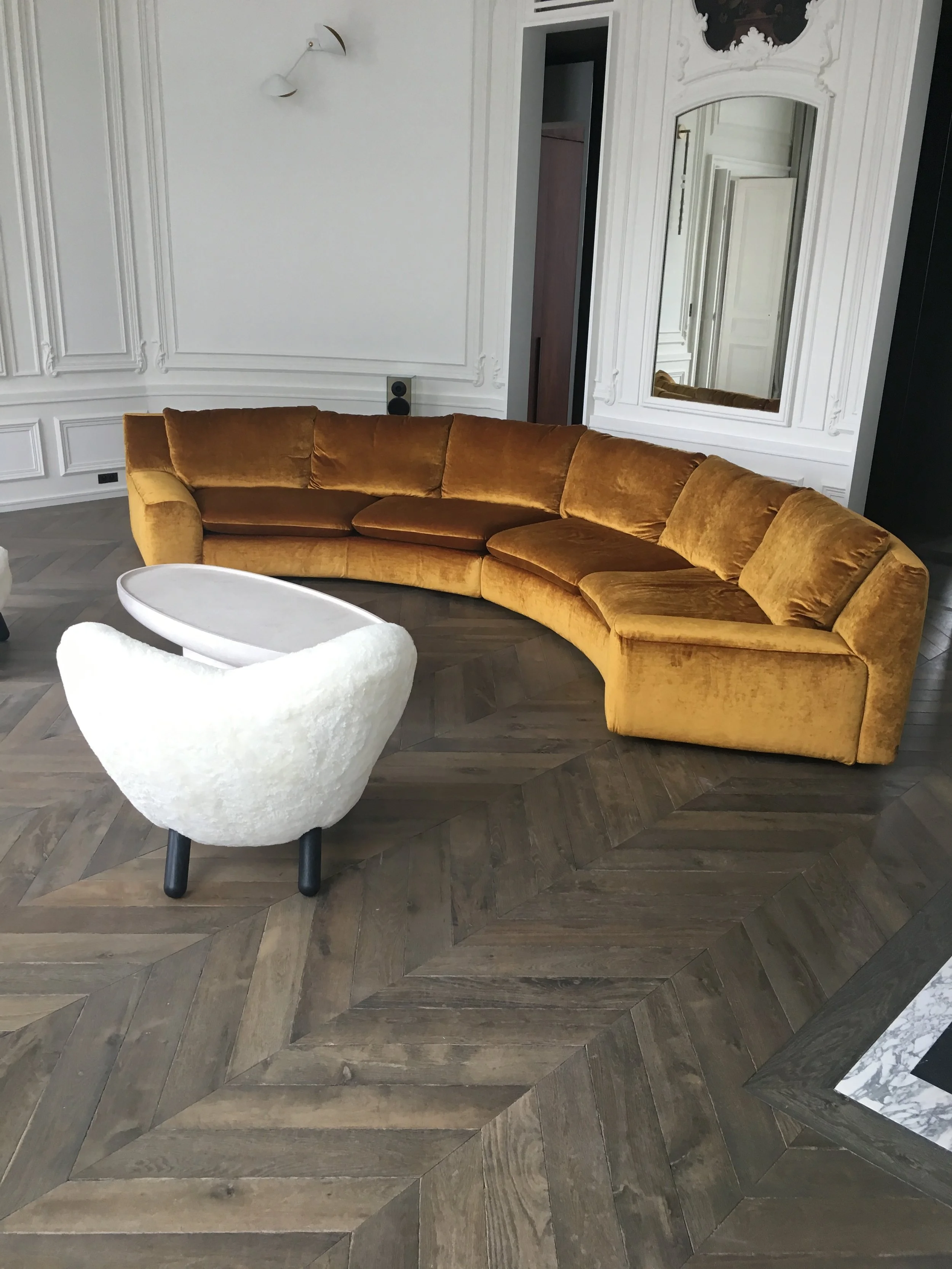 Sala con sofá curvo de terciopelo dorado, silla blanca con patas negras y un espejo grande en la pared con detalles decorativos en blanco y paredes blancas con molduras.