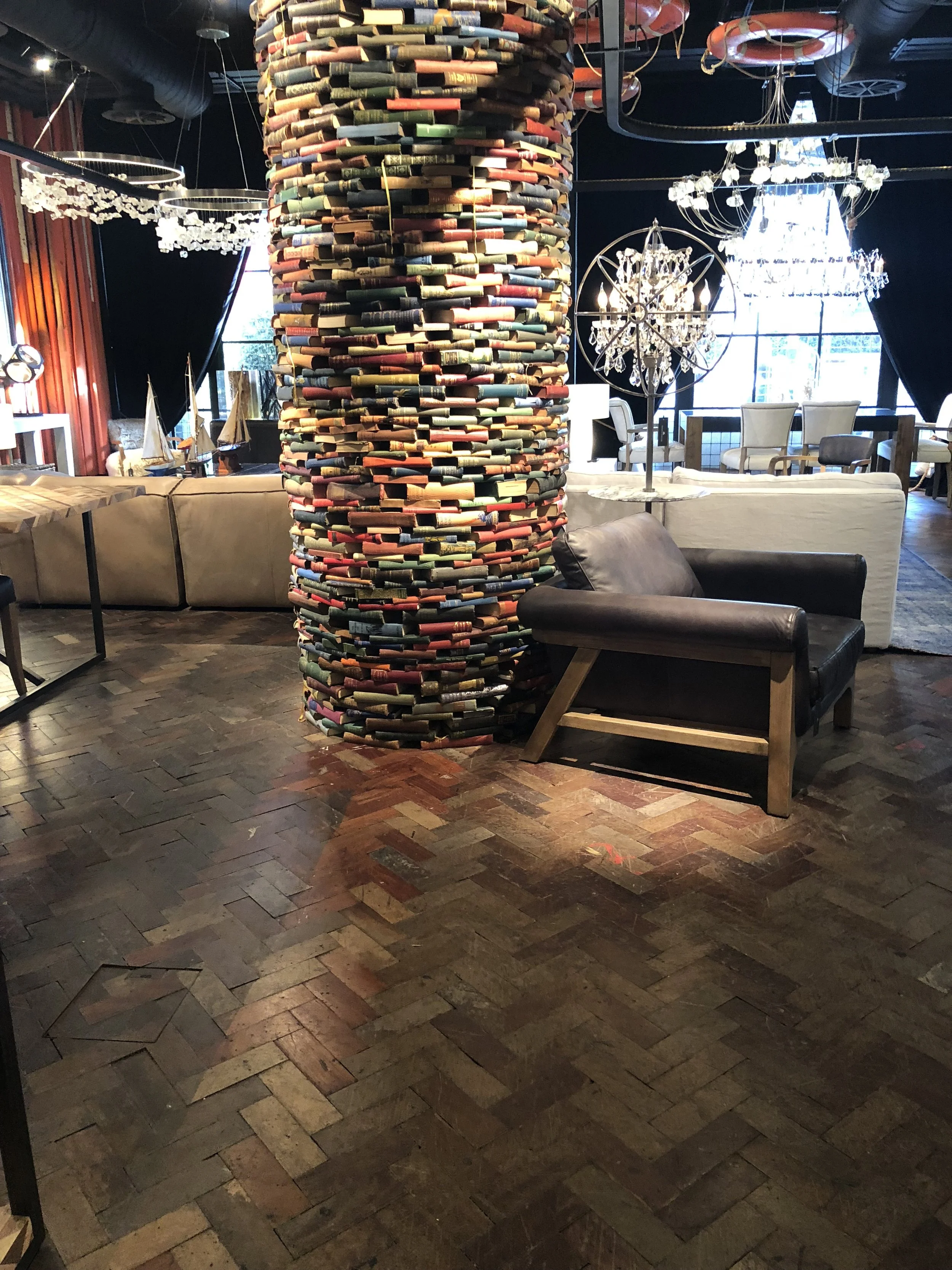 Área de interior con techo oscuro, luminarias decorativas de cristal, muebles de colores neutros, columna decorativa cubierta con libros apilados, y piso de madera en patrón de espiga.