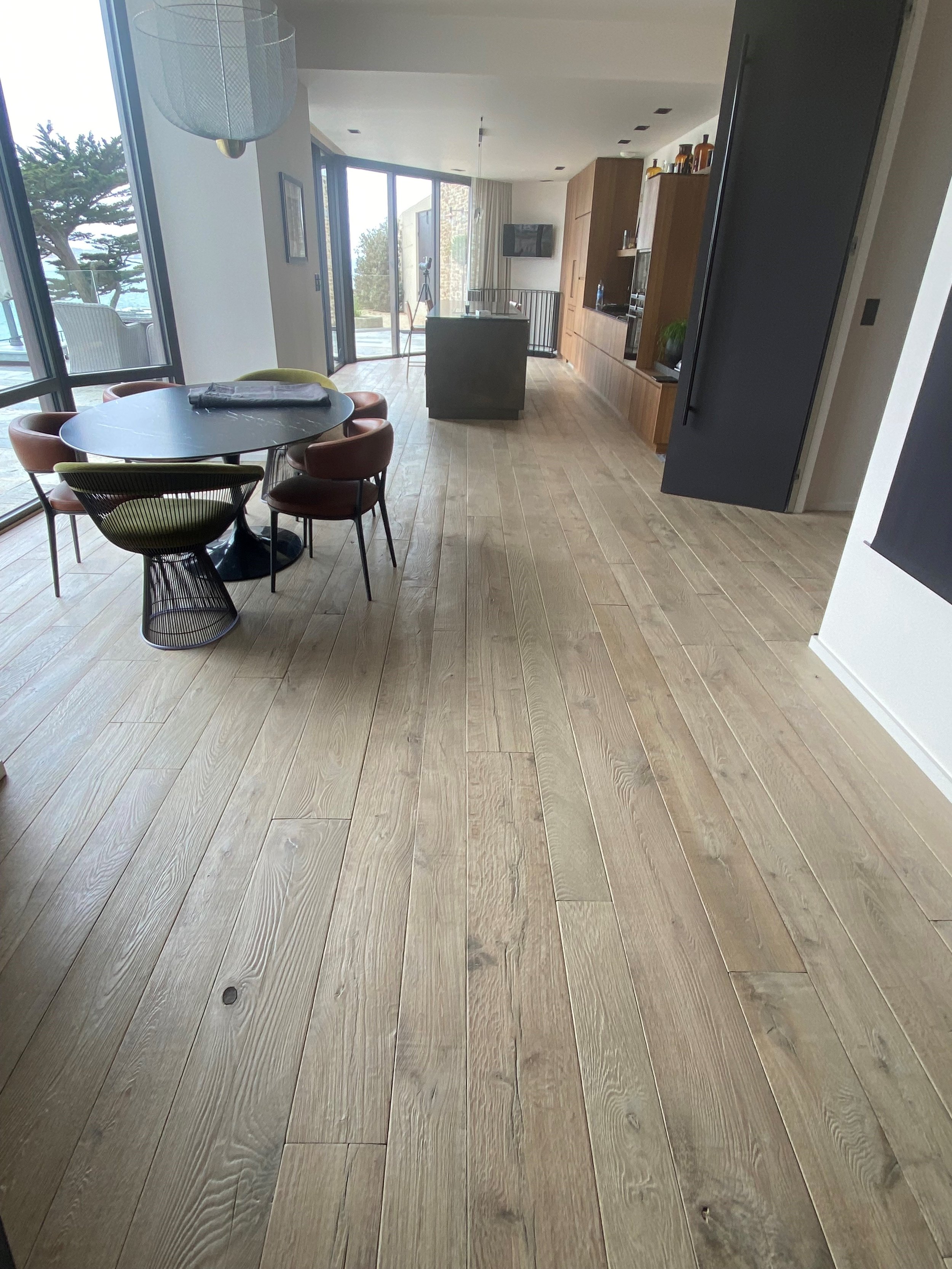 Interior de una sala moderna con piso de madera, una mesa redonda con sillas de diferentes colores, ventanas grandes con vista al exterior, y una cocina al fondo.