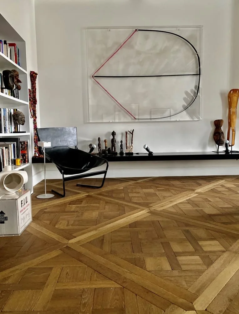 Sala con mueble negro, silla negra, estantería blanca con libros y objetos decorativos, cuadro con línea roja y negra en la pared, piso de madera con patrón diagonal, y varias esculturas en la repisa.