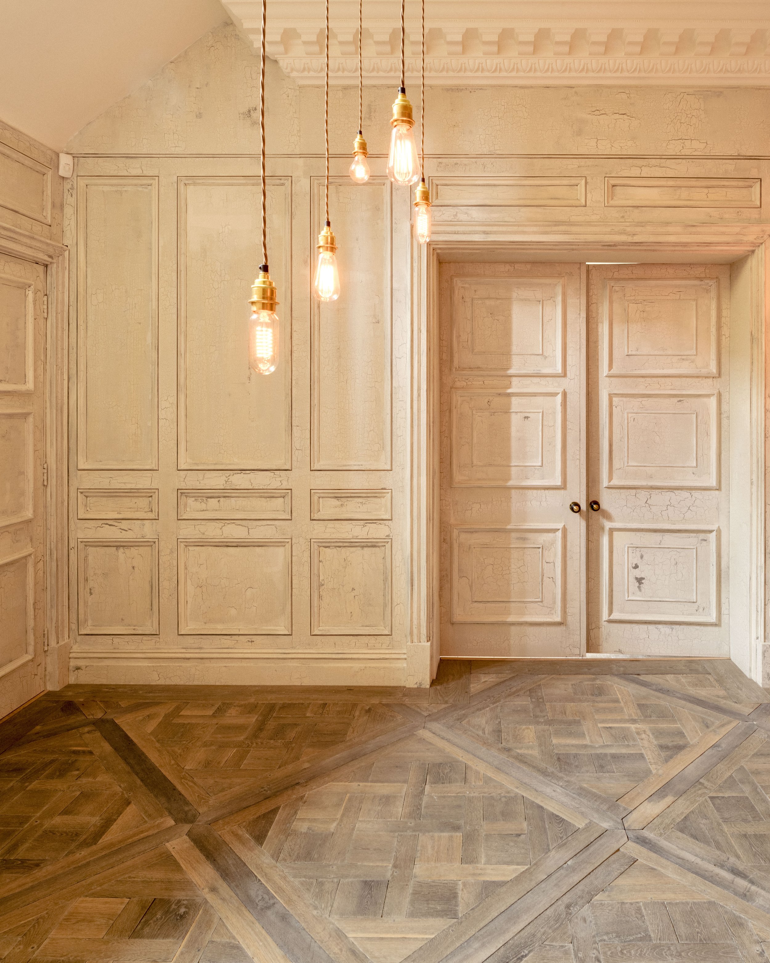 Sala con paredes de color crema, puertas de madera pintadas de blanco con paneles decorativos, y lámparas colgantes con bombillas expuestas.