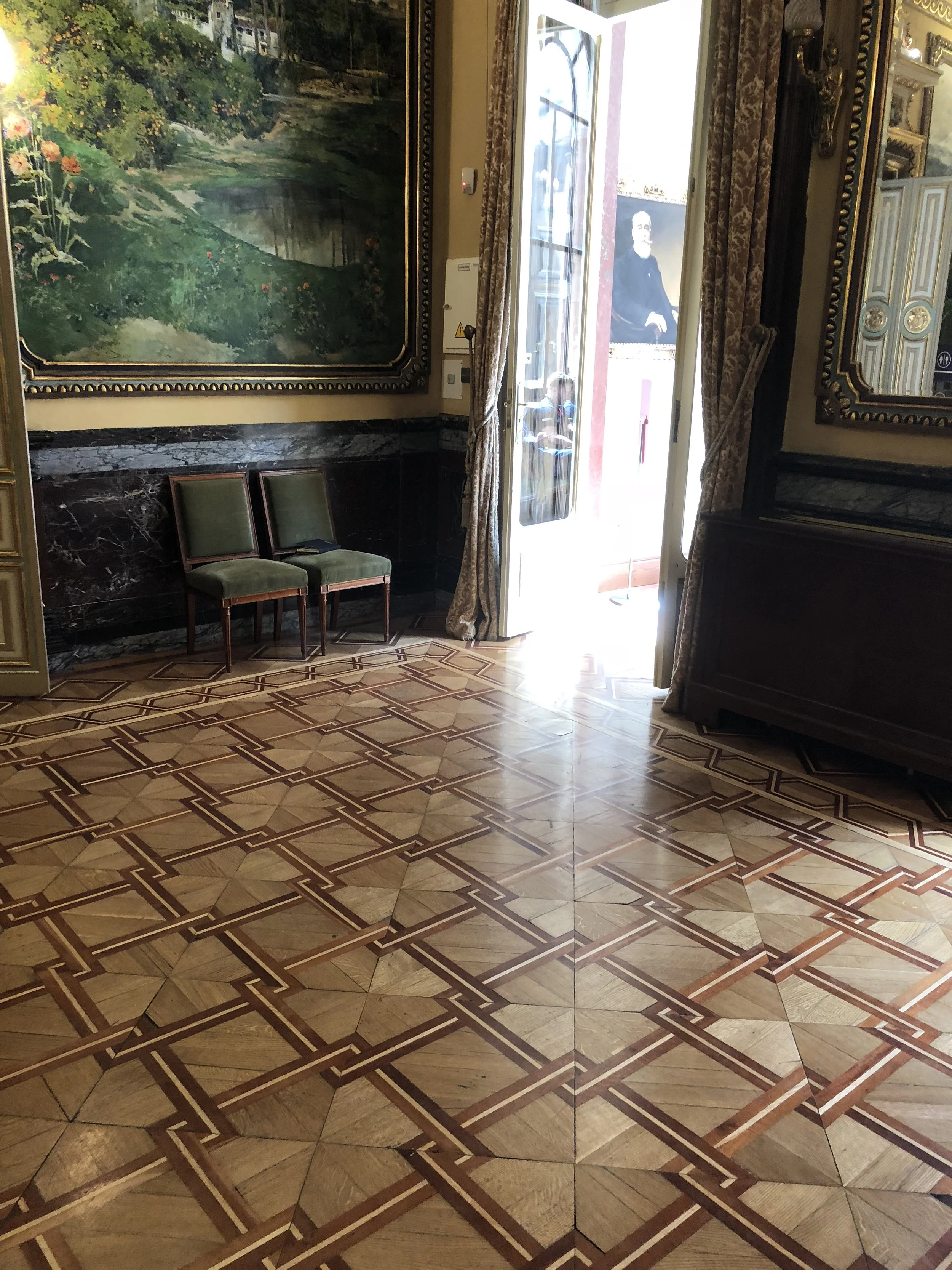 Sala lujosa con piso de madera con diseño geométrico, dos sillas verdes junto a la pared, ventanas con cortinas doradas, pintura grande en la pared y retrato visible en la entrada, luz brillante entrando por la puerta abierta.