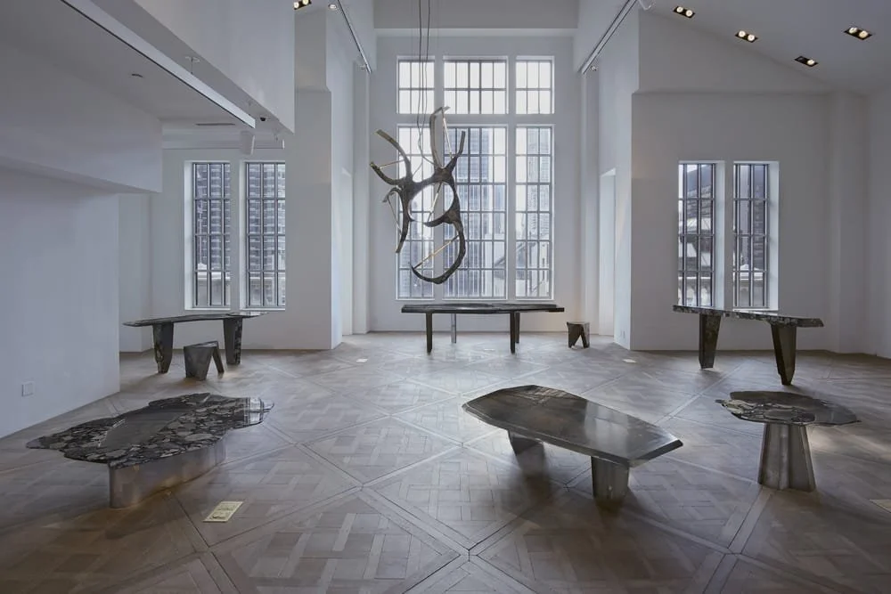 Sala de exposición con mesas y una escultura de cuernos en la pared, ventanas grandes y piso de madera.