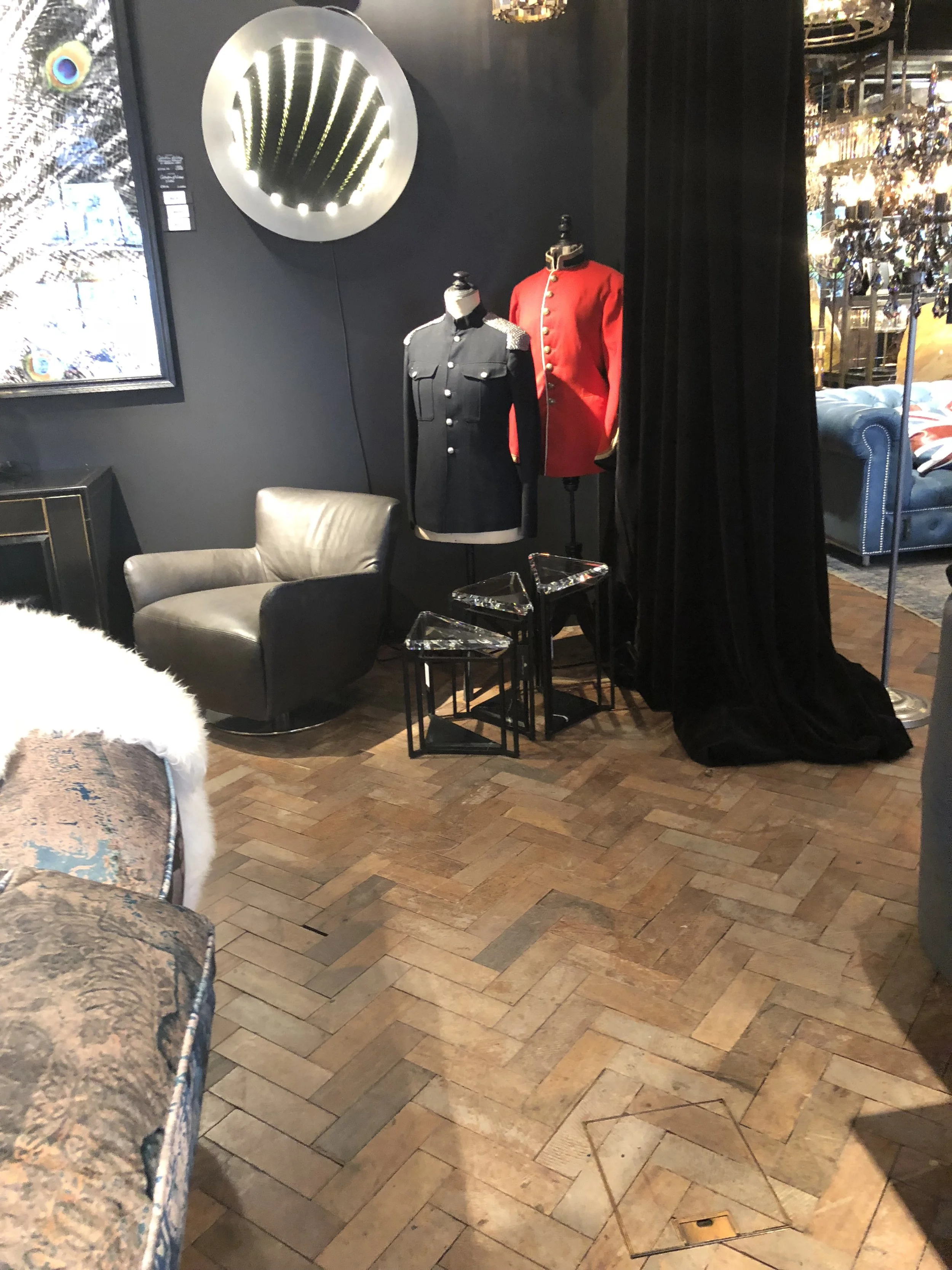 Dos maniquíes con uniformes militares, uno negro y uno rojo, en una sala con muebles modernos y un espejo redondo con luces.
