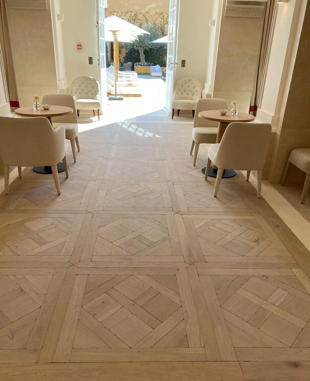 Interior de un salón con sillas y mesas redondas decoradas con pequeñas flores, y puertas abiertas que muestran un área exterior con sombrillas y tumbonas.