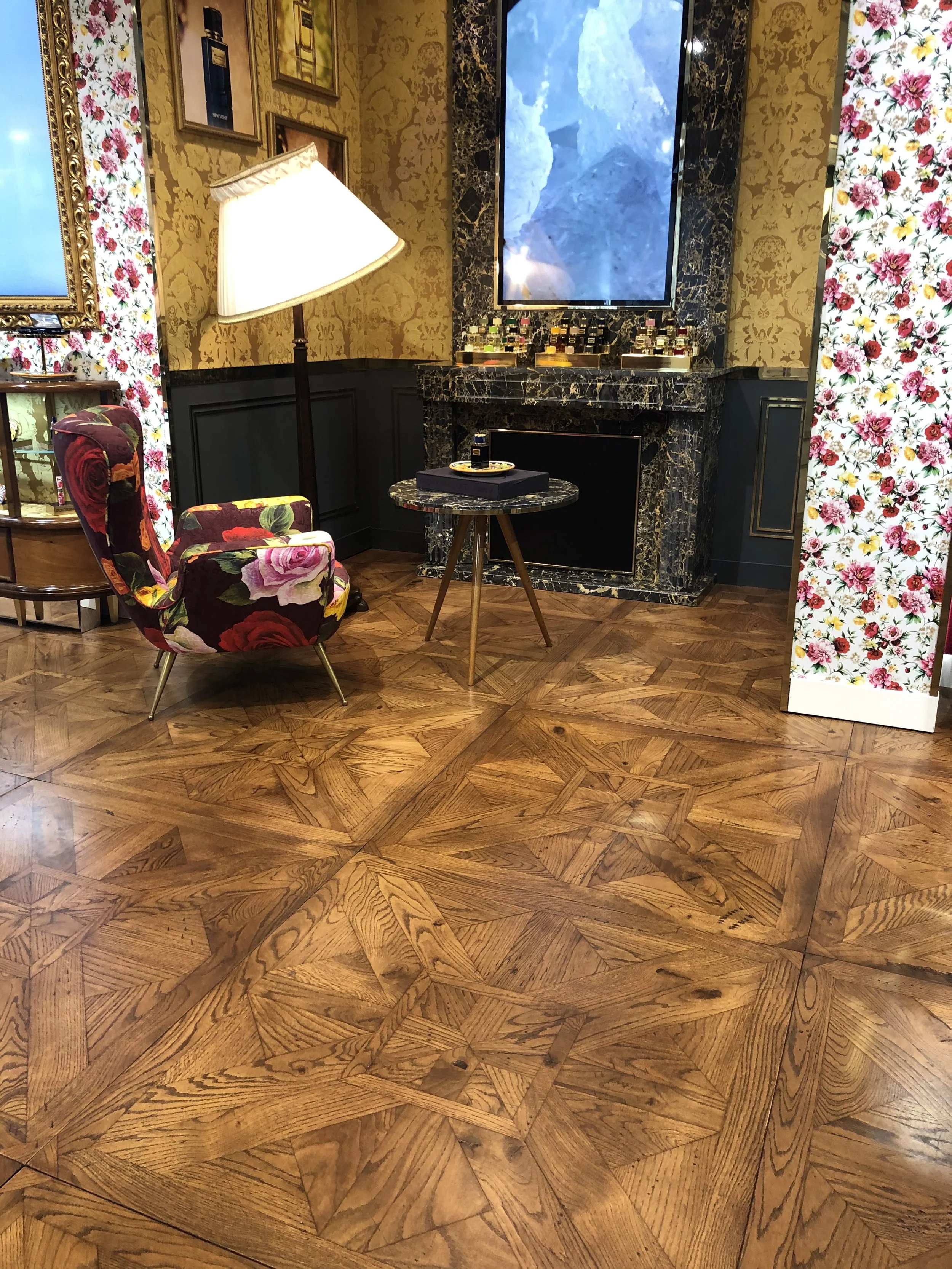 Interior decorativo con una silla tapizada con tela de flores, una lámpara de pie, una mesa redonda pequeña, un marco de fotos en la pared, un mueble de madera y una pantalla de televisión. El suelo es de madera con un patrón geométrico.