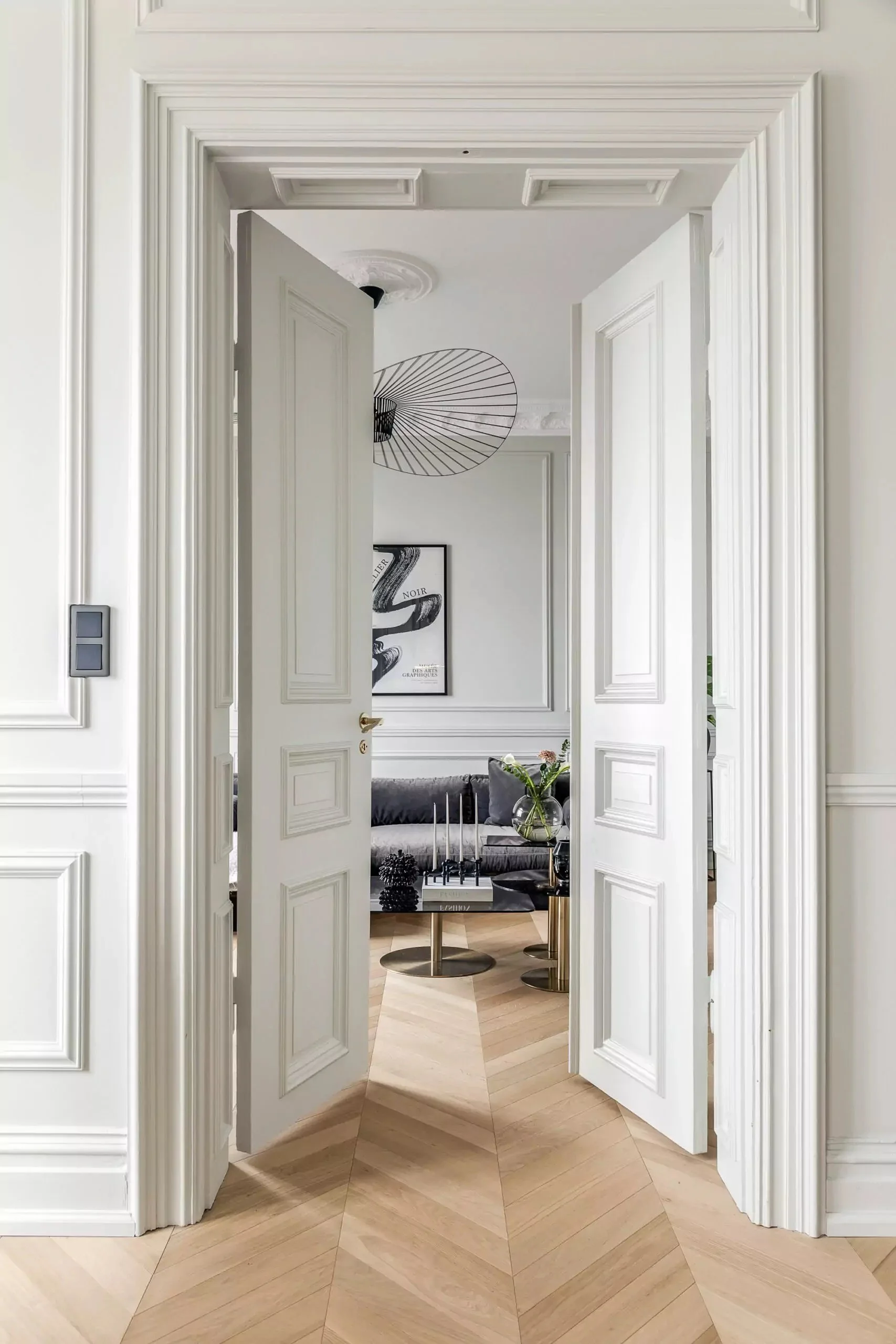 Vista de una sala moderna con paredes blancas con molduras, un sofá gris, mesa de centro negra con objetos decorativos y una pared con arte abstracto enmarcado, vista a través de una puerta doble abierta en un entorno elegante y minimalista.