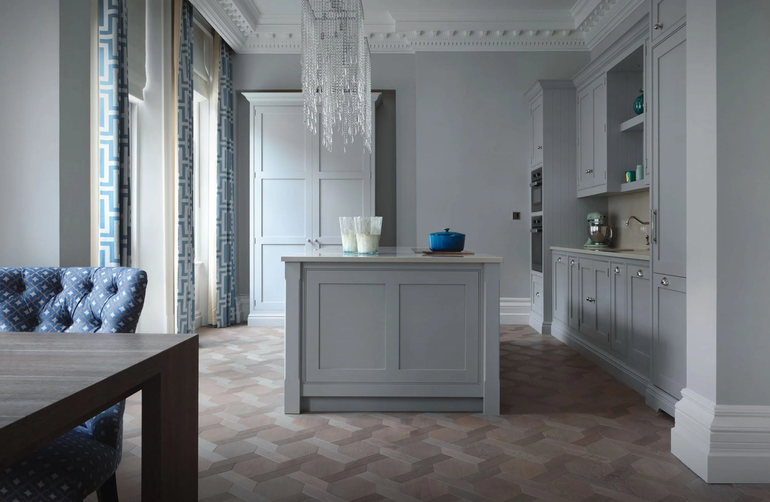 Cocina moderna con gabinetes grises, isla central, lámpara de cristal y cortinas azules con patrones blancos