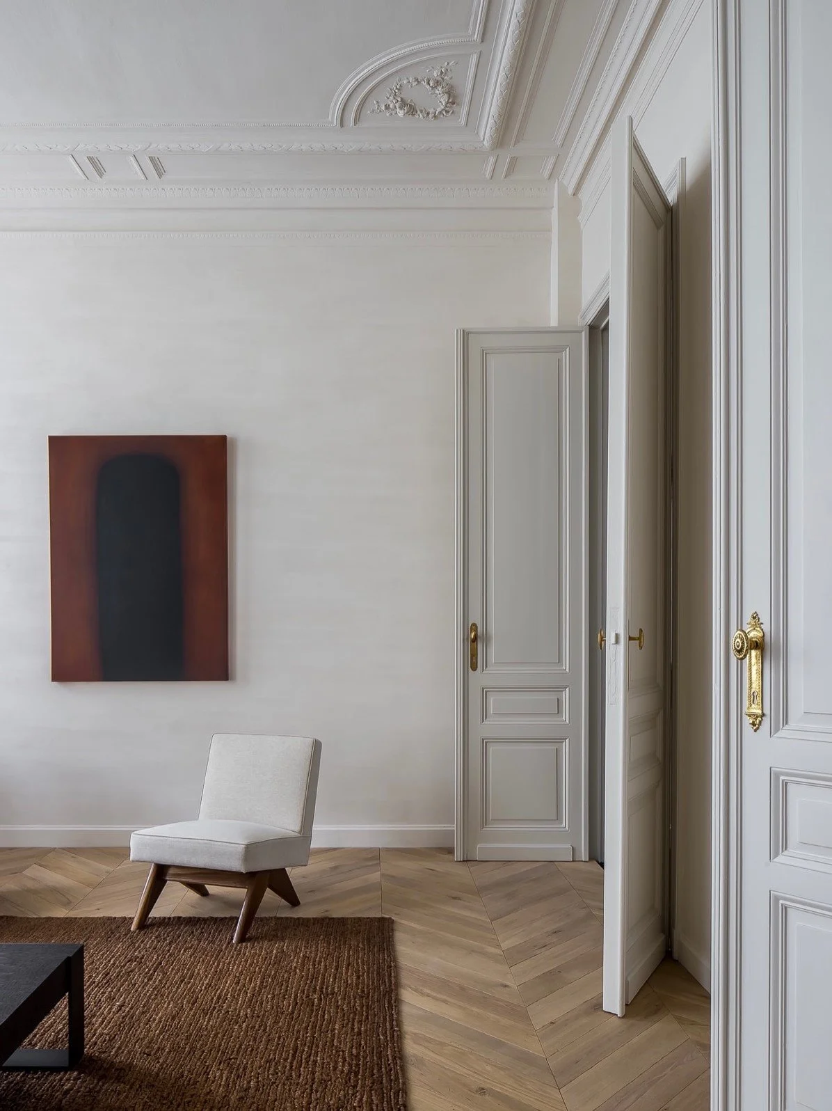 Sala elegante con pared blanca, cuadro abstracto, silla beige, alfombra marrón, puertas blancas con detalles dorados y molduras decorativas en el techo.