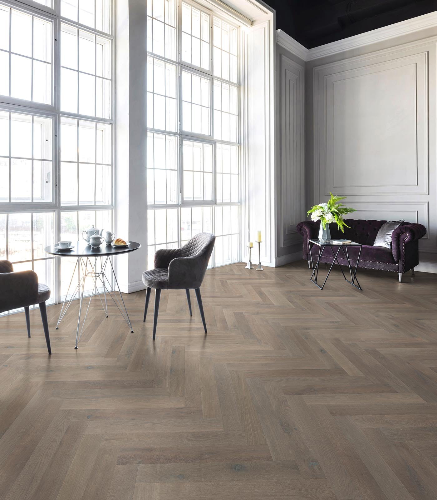 Sala moderna con grandes ventanas de cristal, una mesa redonda con taza, cafetera y croissant, dos sillas y un sofá lila con una mesa con una jarra de flores y un libro.