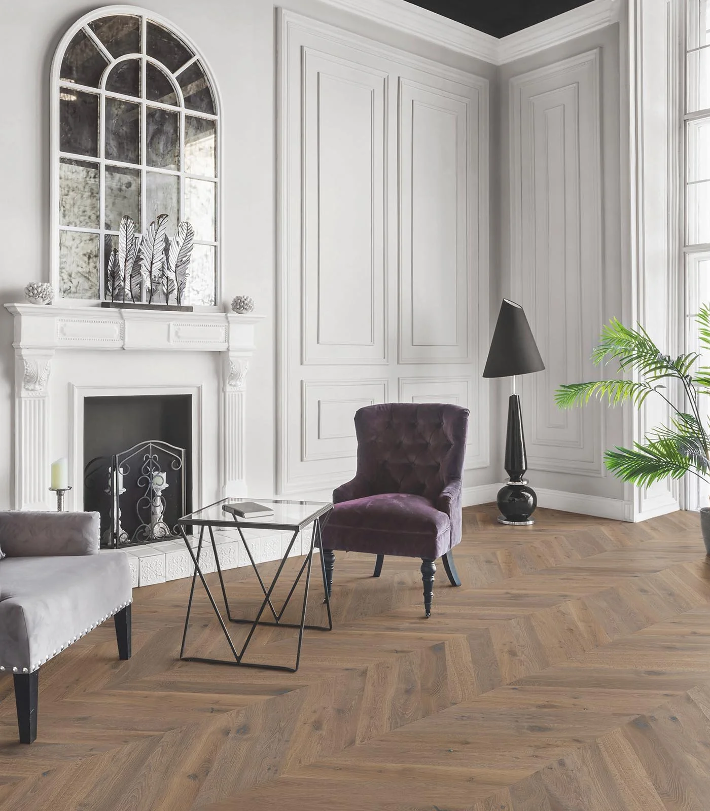 Sala de estar elegante con chimenea blanca, sillón de terciopelo púrpura, lámpara negra y planta decorativa en una habitación luminosa con grandes ventanas y suelos de madera en patrón de espiga tipo chevron en punta Hungría en madera de roble francé