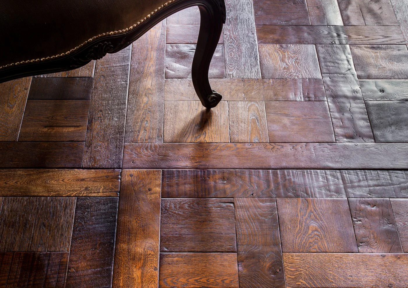 Parte de una silla de madera oscura con detalles tallados y clavos de oro en la orilla, sobre un piso de madera pulida con patrones de listones rectos de diferentes tonos.