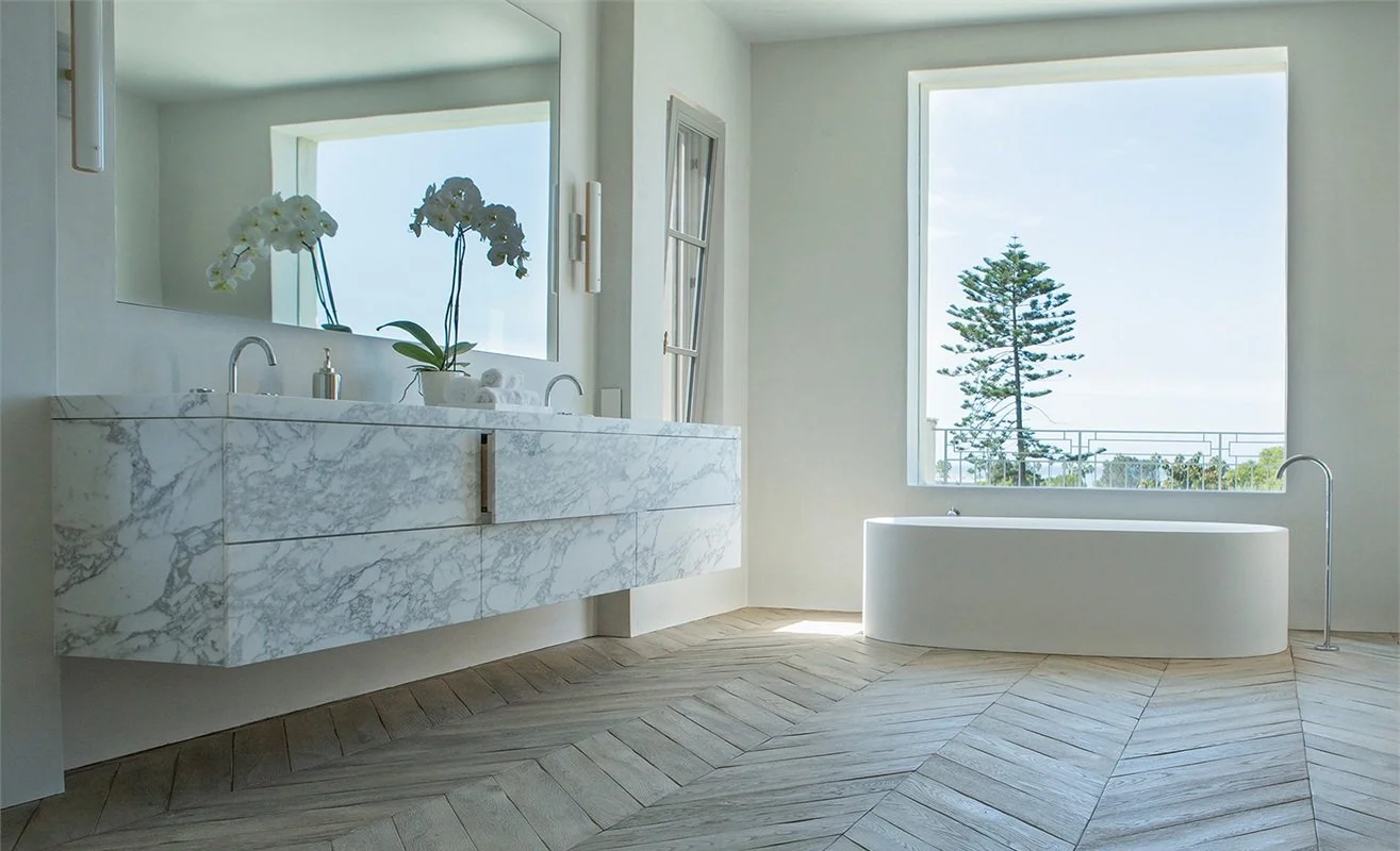 Baño con lavabo de mármol, espejo grande, bañera con vista a una ventana con vista a un árbol y el cielo, decoración minimalista con plantas en el lavabo.