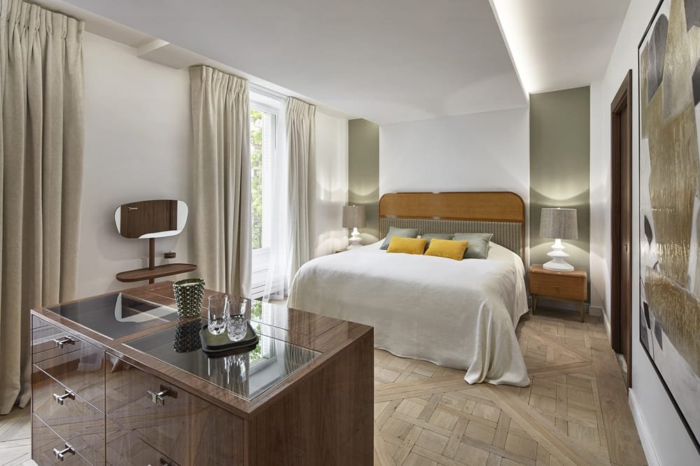 habitación de hotel con cama grande, cabecera de madera, dos lámparas, mesitas, cortinas beige, escritorio con botella y vasos, una ventana con vista a árboles.