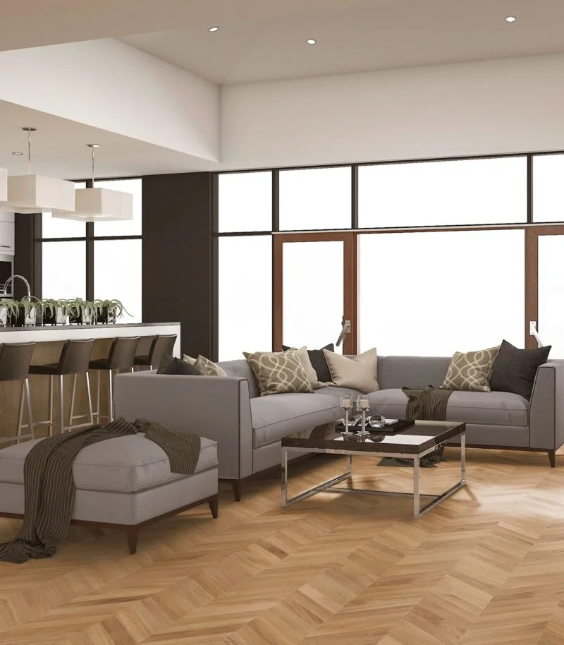 Sala moderna con sillón y ottoman gris, mesa de centro y cojines decorativos, en un espacio con grandes ventanas y suelos parquet de madera.