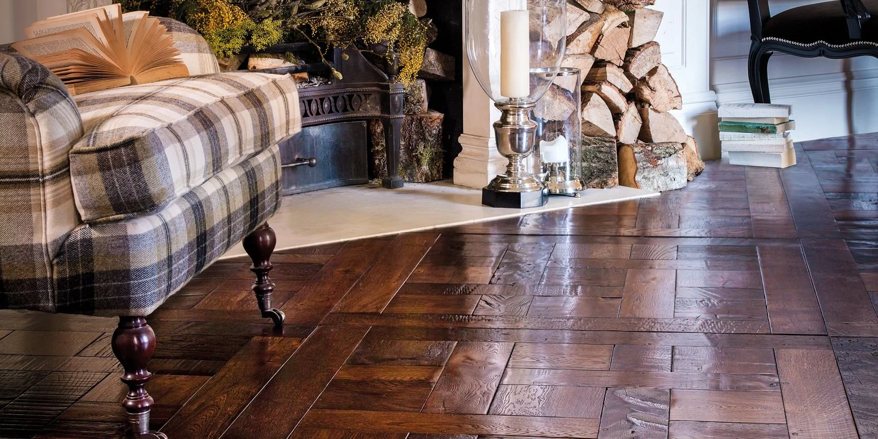 Sala de estar con chimenea con leña apilada, una alfombra sobre pisos de madera pulida, una lámpara con candelabro, varias pilas de libros apilados y un sofá con tapizado de tartán.