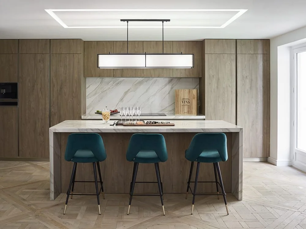 Cocina moderna de madera clara con isla de mármol, tres sillas azules y una lámpara rectangular colgante.