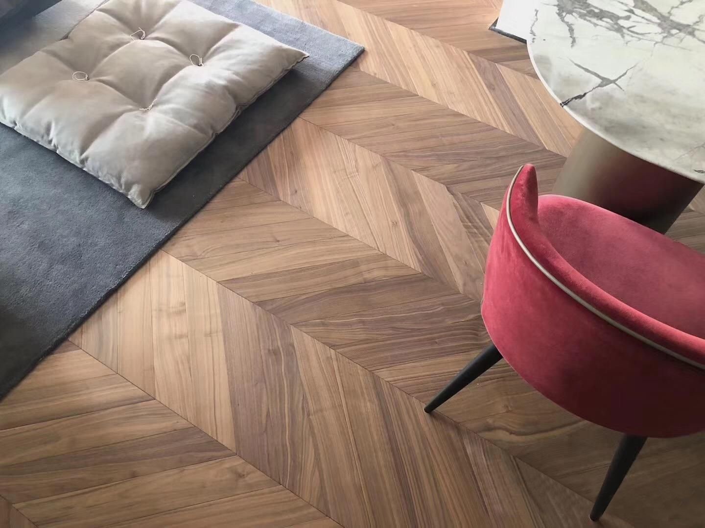 Suelo de madera con patrón en espiga, alfombra gris y cojín beige en el suelo, junto a una mesa de mármol y una silla de terciopelo roja