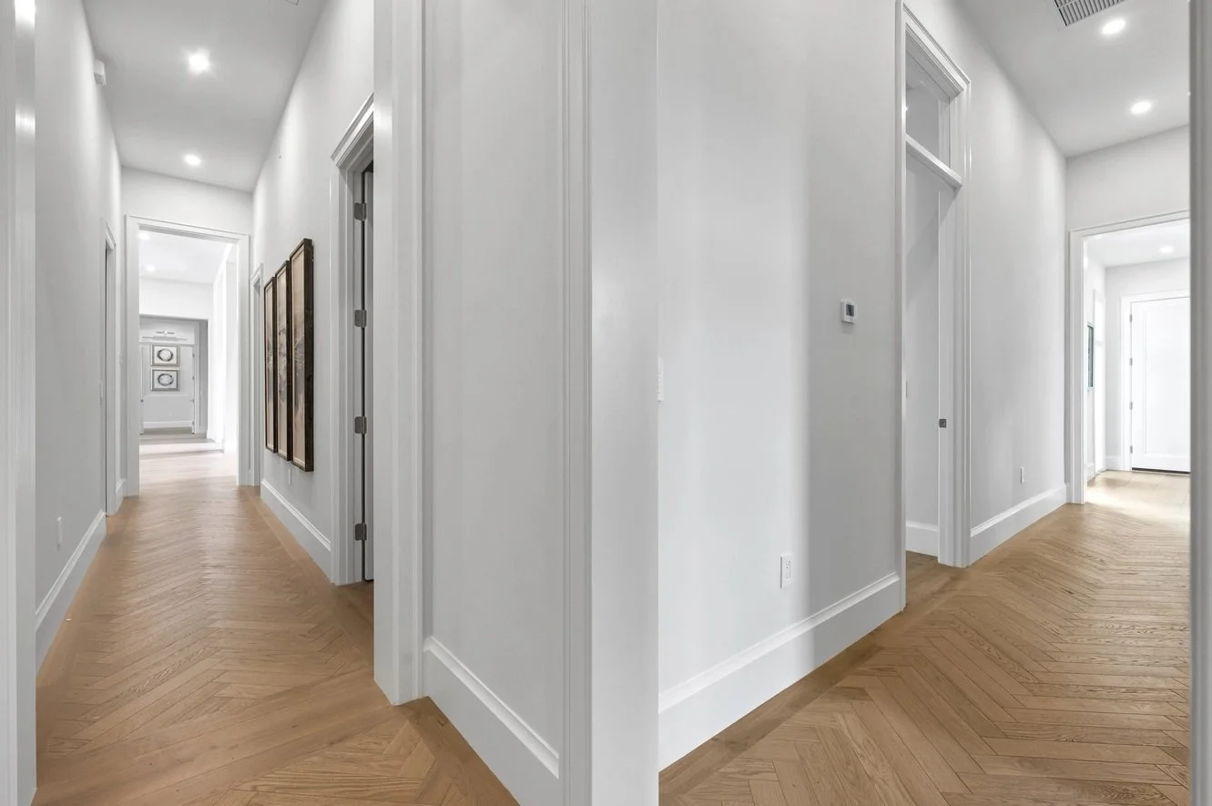 Pasillo de una casa con paredes blancas, piso de madera en espiga, varias puertas y cuadros decorativos en las paredes.