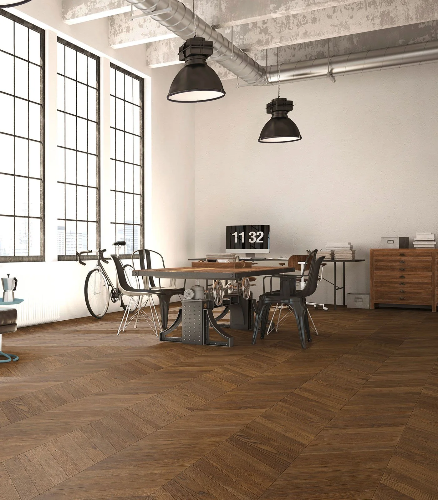 Oficina moderna con grandes ventanas de cristal, iluminación industrial, escritorio redondo con varias sillas, bicicleta apoyada en la pared, escritorio secundario con archivos y una pantalla en la pared mostrando la hora 11:32.