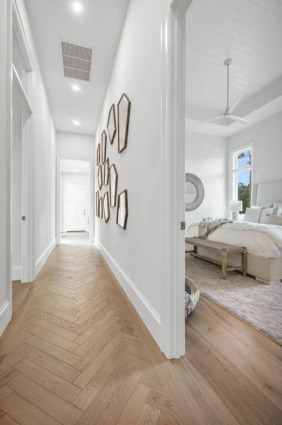 Pasillo con paredes blancas, suelo de madera en patrón de espiga, y decoraciones de cuadros en la pared. A la derecha, vista de una habitación con cama y ventanas que dejan entrar luz natural.