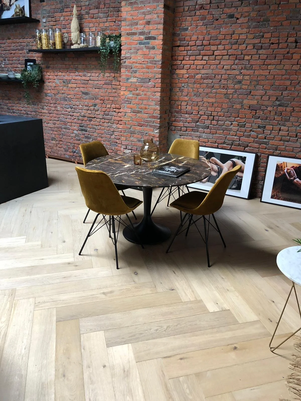 Mesa redonda de mármol con cuatro sillas de respaldo en color mostaza en una habitación con paredes de ladrillo expuesto y cuadros apoyados en el suelo.