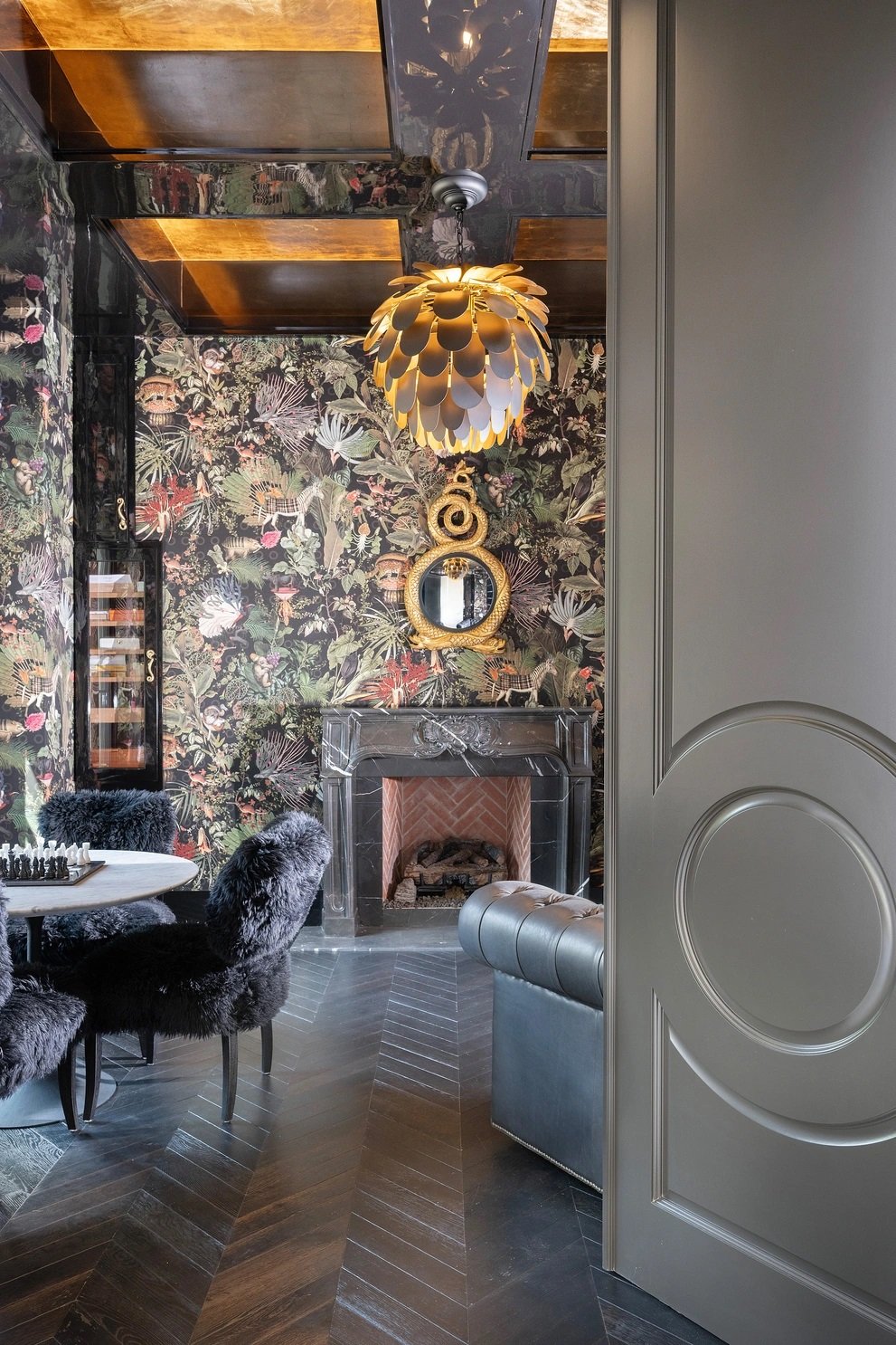 Interior elegante con chimenea de mármol, pared con papel tapiz con diseño floral oscuro, lámpara moderna dorada con pétalos, espejo dorado sobre la chimenea, sillones con cubres de peluche, y una mesa de juego de ajedrez.