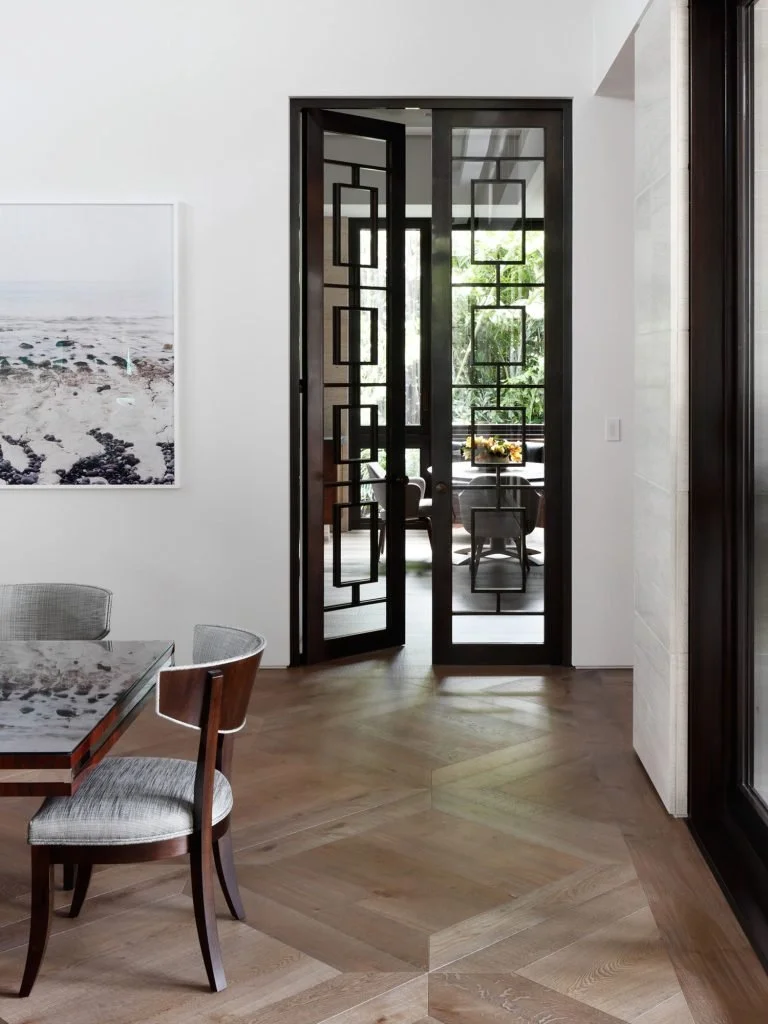 Puerta de vidrio con marco negro y diseño geométrico en una casa moderna, vista hacia una sala con ventanas de grandes dimensiones y muebles en el fondo.