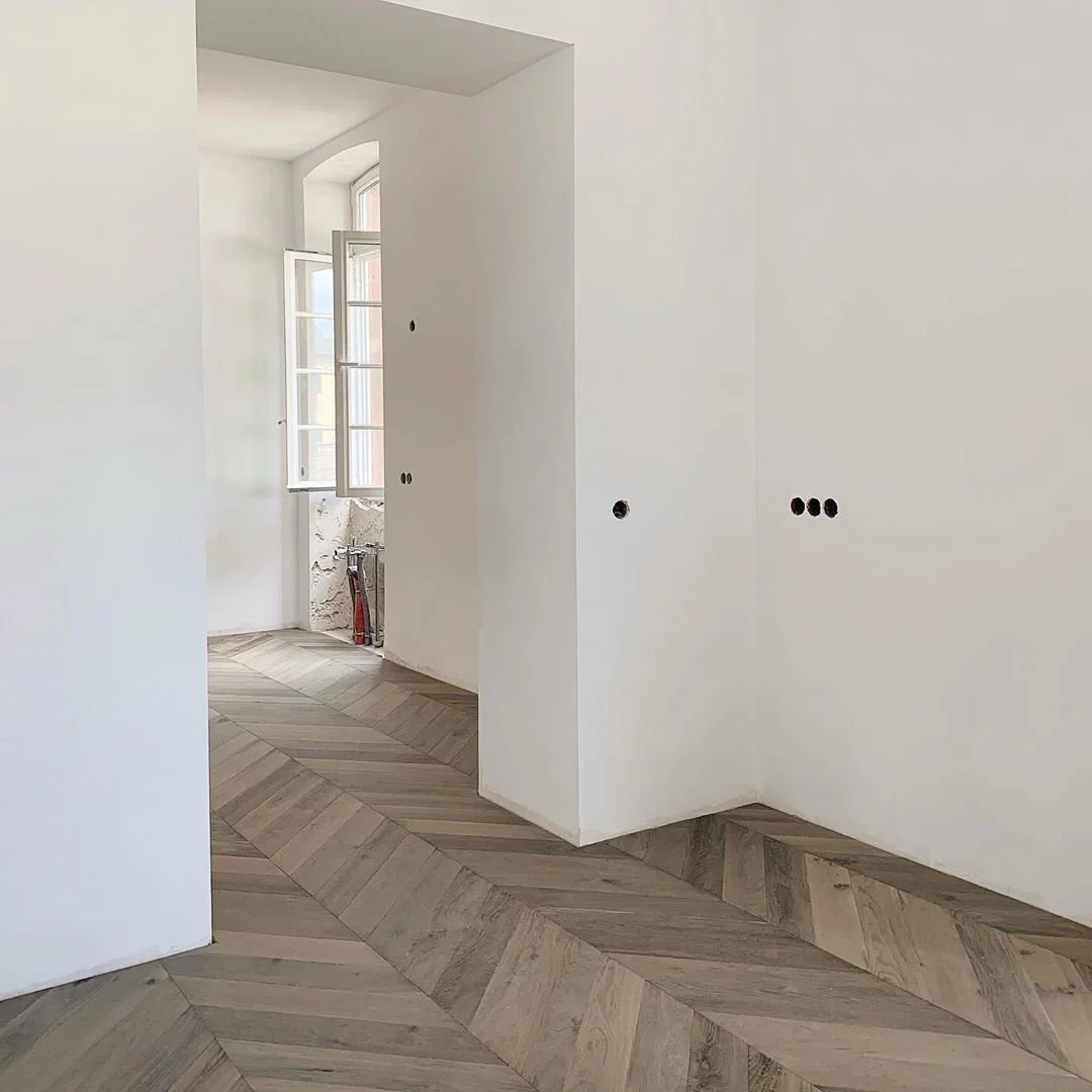 Paredes blancas en proceso de construcción con agujeros para instalaciones eléctricas y una ventana doble abierta que deja entrar luz natural en una habitación con suelos parquet de madera. en patrón diagonal.