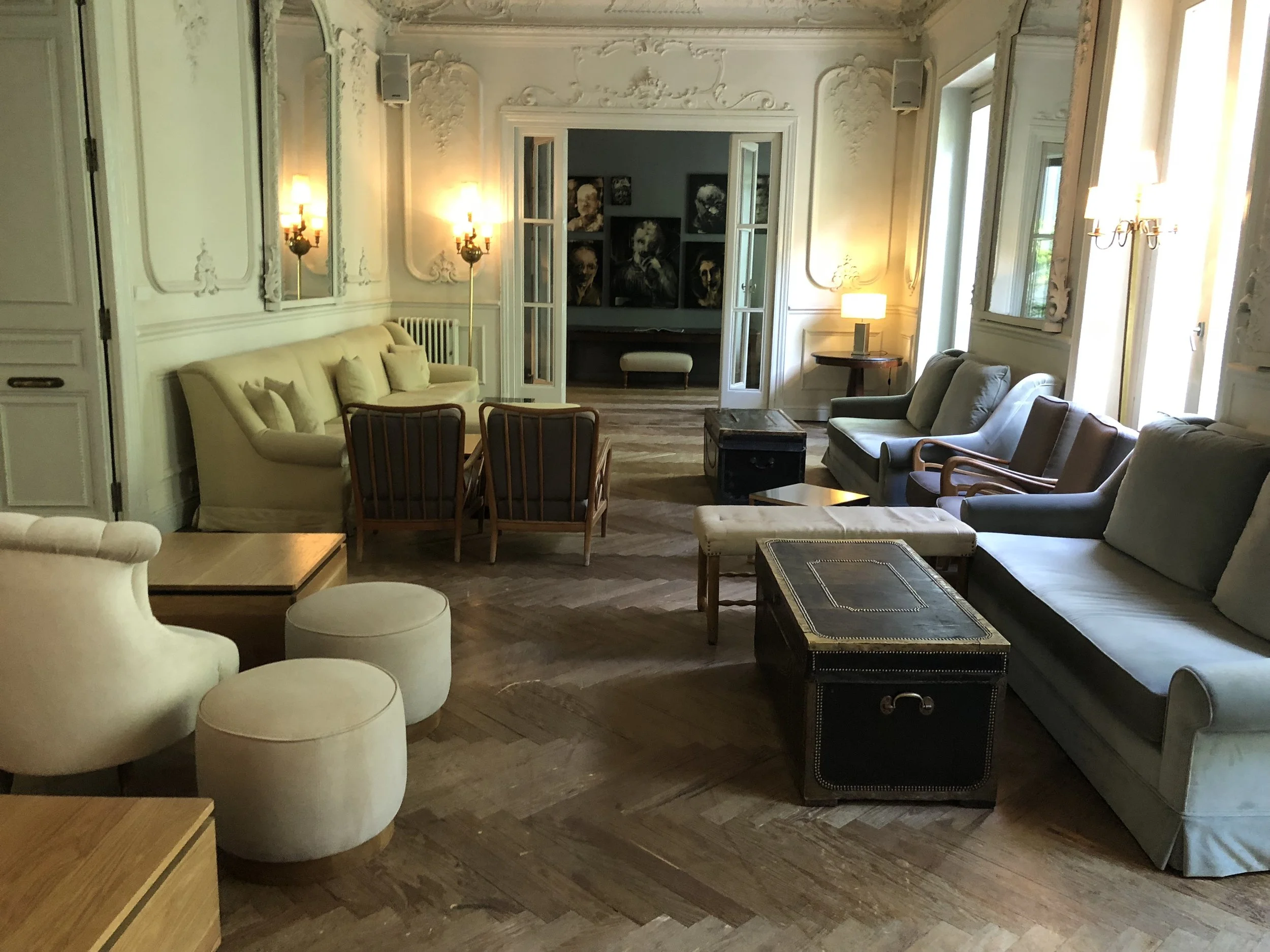 Sala de estar elegante con sillones y sillas de diferentes estilos, mesas de madera y pequeños puffs, con paredes decoradas y luces cálidas.