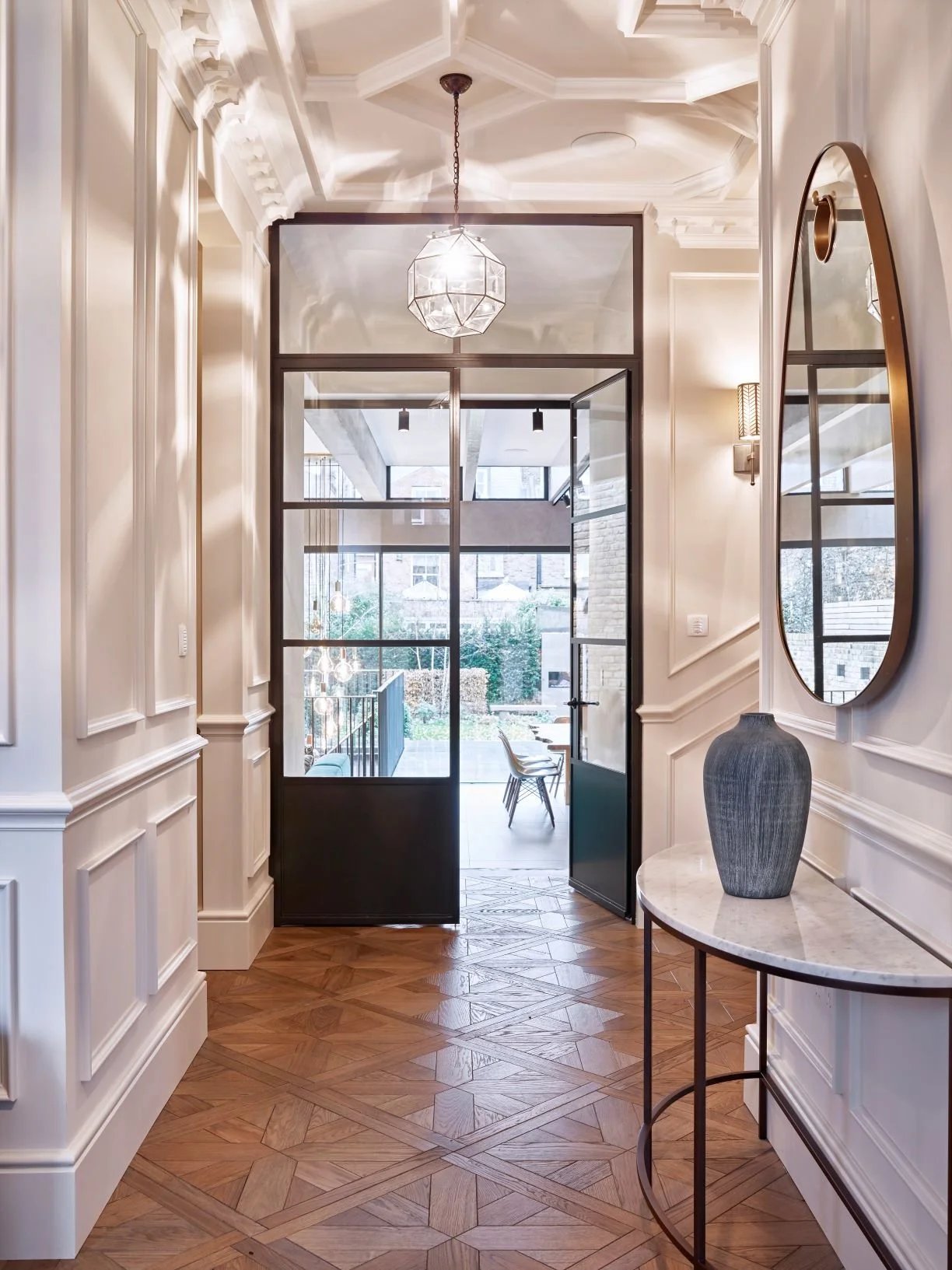 Entrada elegante con paredes blancas decoradas y piso de parquet de madera, con un espejo ovalado y una lámpara moderna en la pared, puerta de vidrio y vistas a un patio con mesas y sillas.