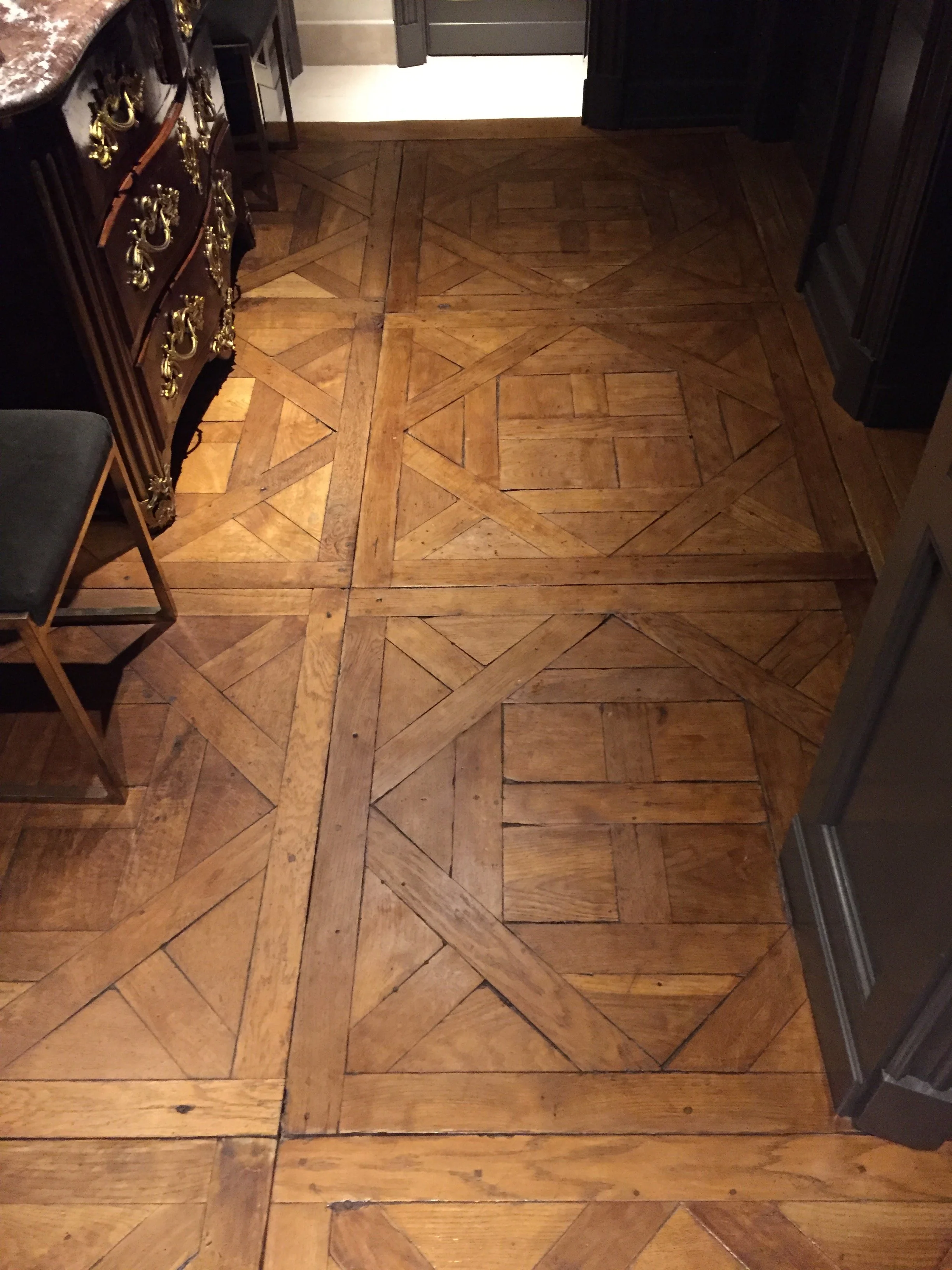 Piso de madera con diseño en cuadros y líneas rectas, en un ambiente interior con muebles oscuros y una silla de madera con cojín gris.