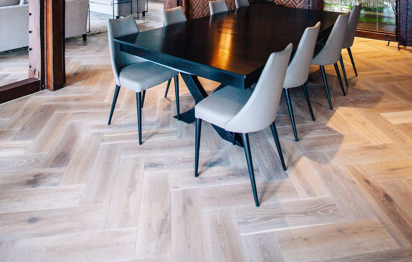 Mesa negra rectangular con sillas grises alrededor en una sala moderna con suelo de madera en patrón herringbone.