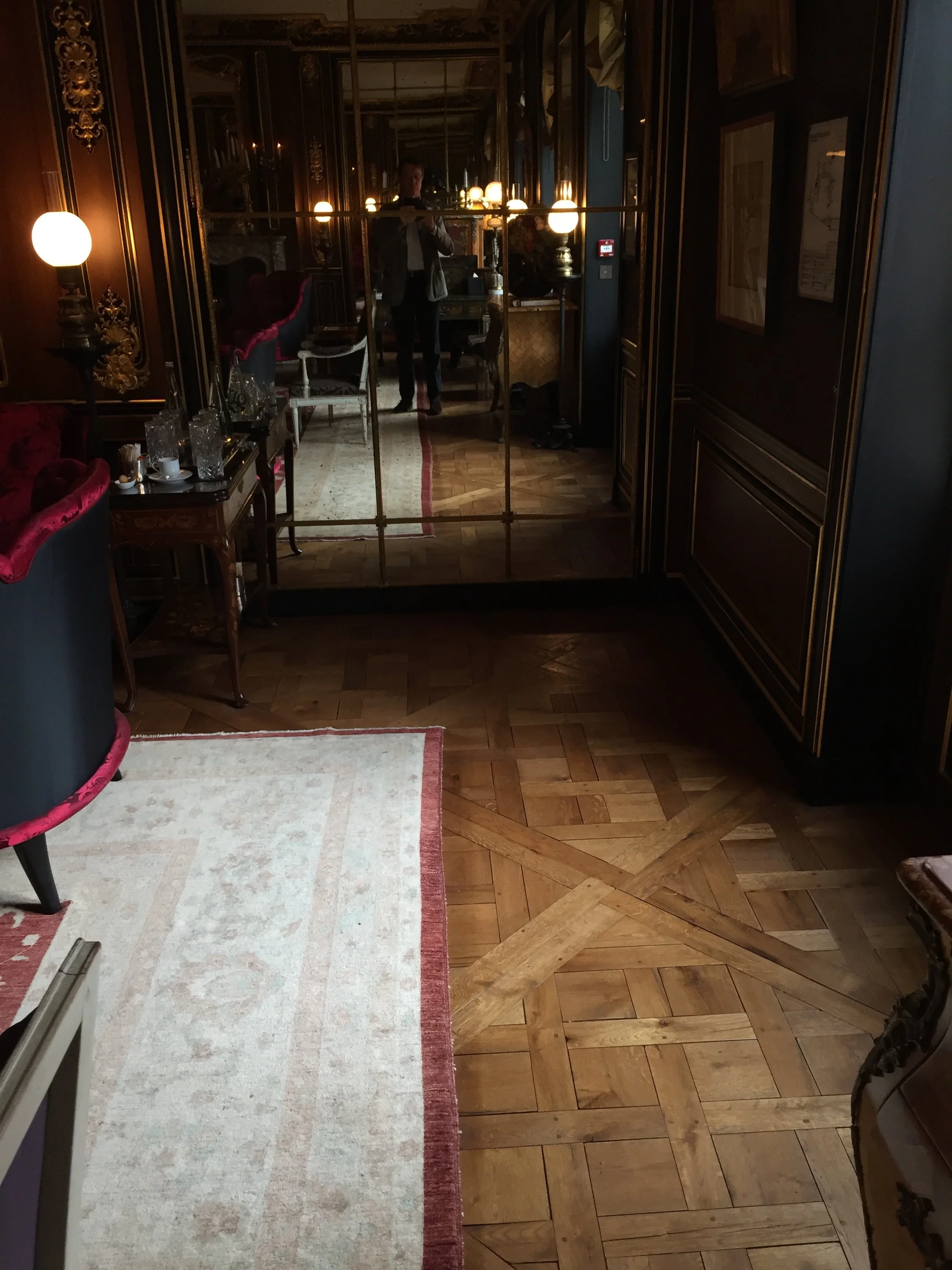 Reflejo en un espejo grande que muestra una sala decorada con estilo clásico, con muebles antiguos, lámparas de брос, paredes con molduras y cuadros. Se puede ver a una persona tomando la foto y una alfombra en el suelo de madera.