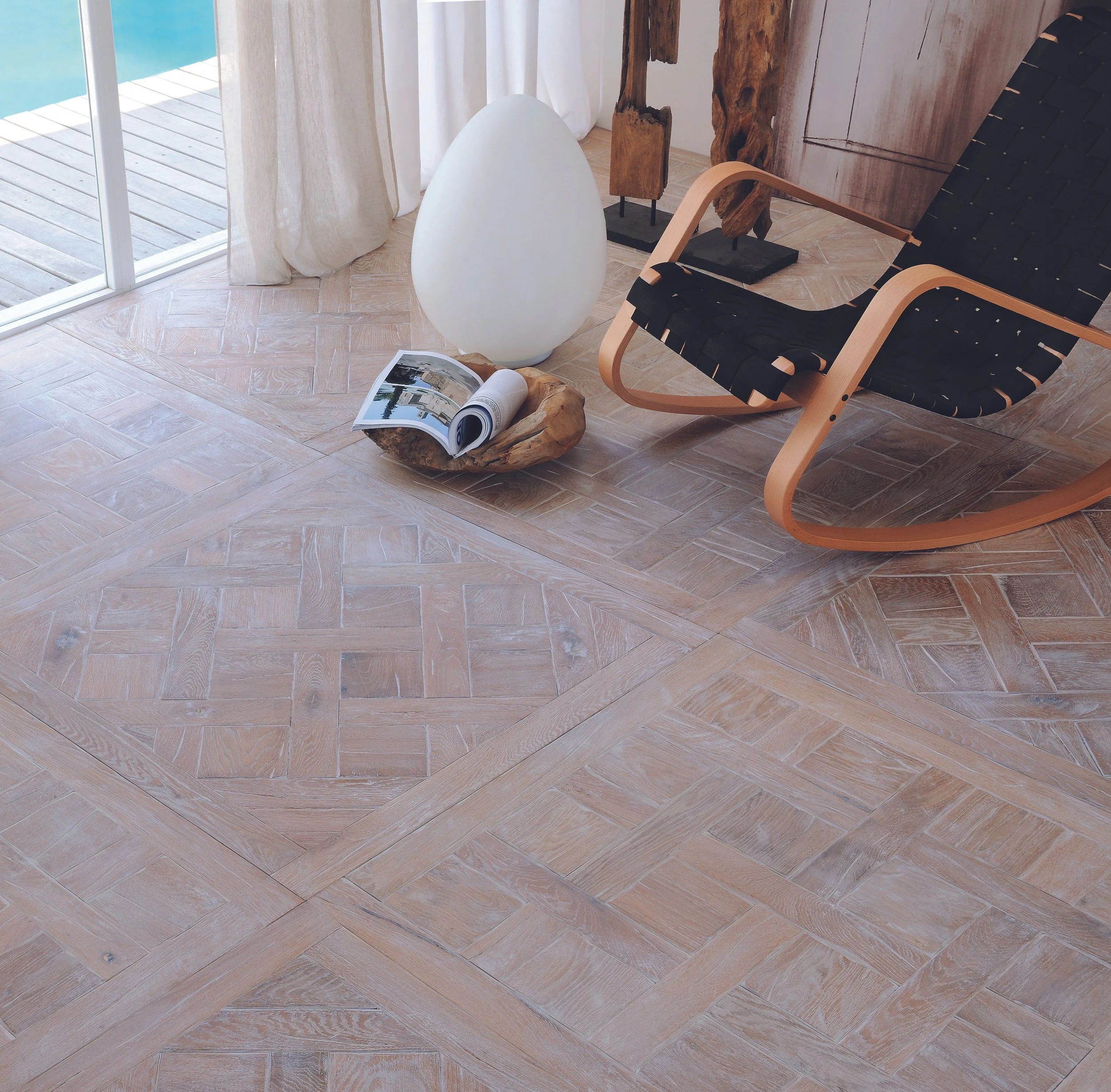 Rincón de interior con piso de madera, silla de madera y cuerda, lámpara de pie blanca, ventana con cortinas, y un libro abierto sobre una pieza de madera.