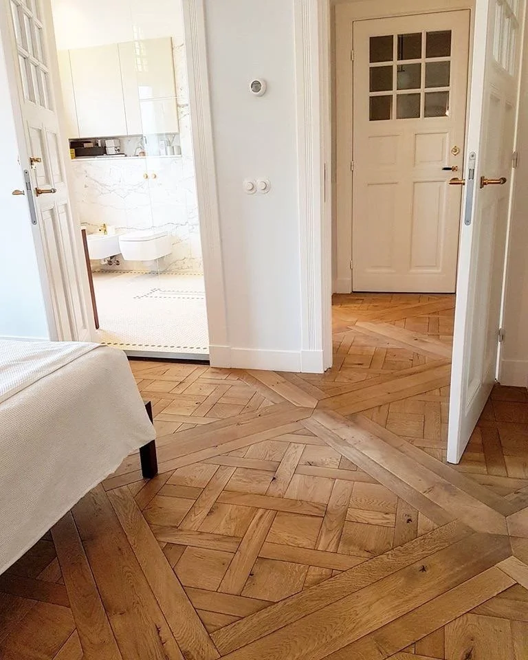 Vista de una sala y un baño con paredes blancas, puertas blancas y pisos de madera. Puerta del baño abierta mostrando lavabo y azulejos blancos.