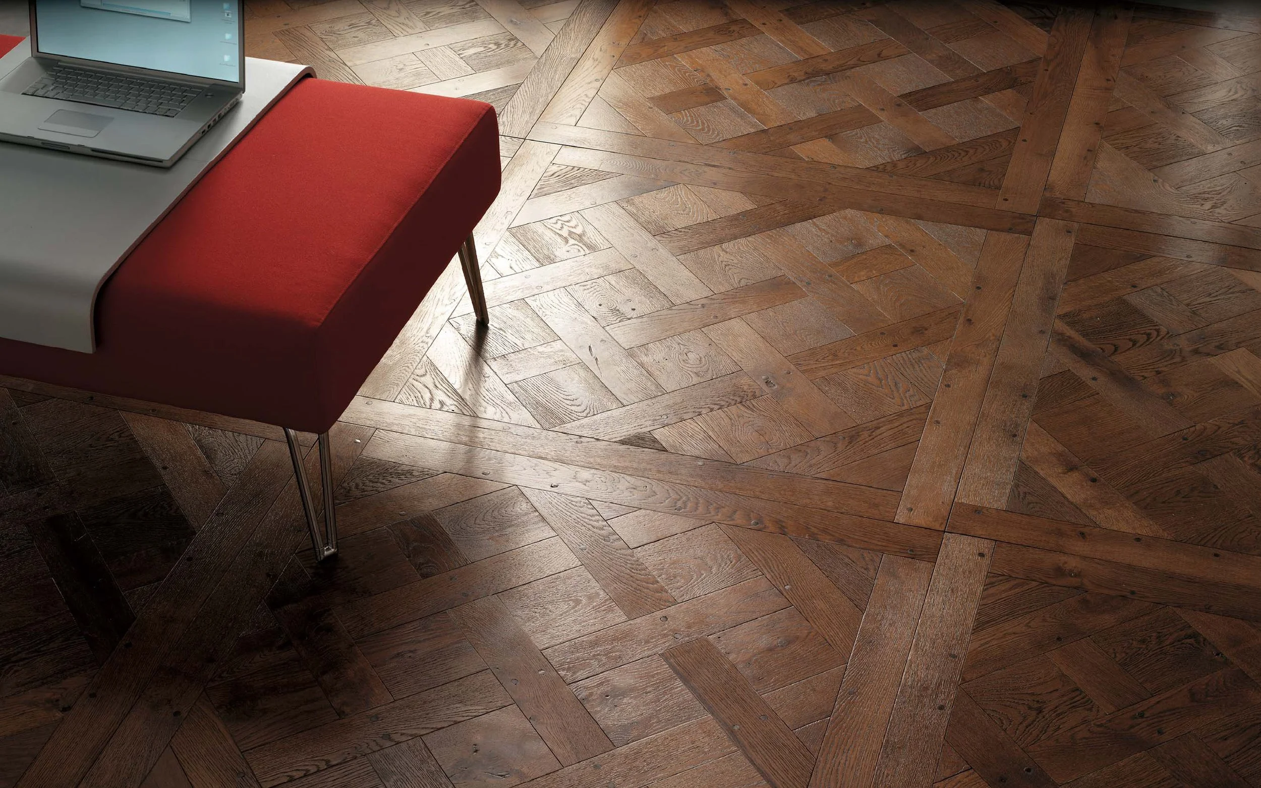 Madera pulida con patrón entrelazado en un piso de interior, muestra una mesa con cojín rojo en la esquina superior izquierda.